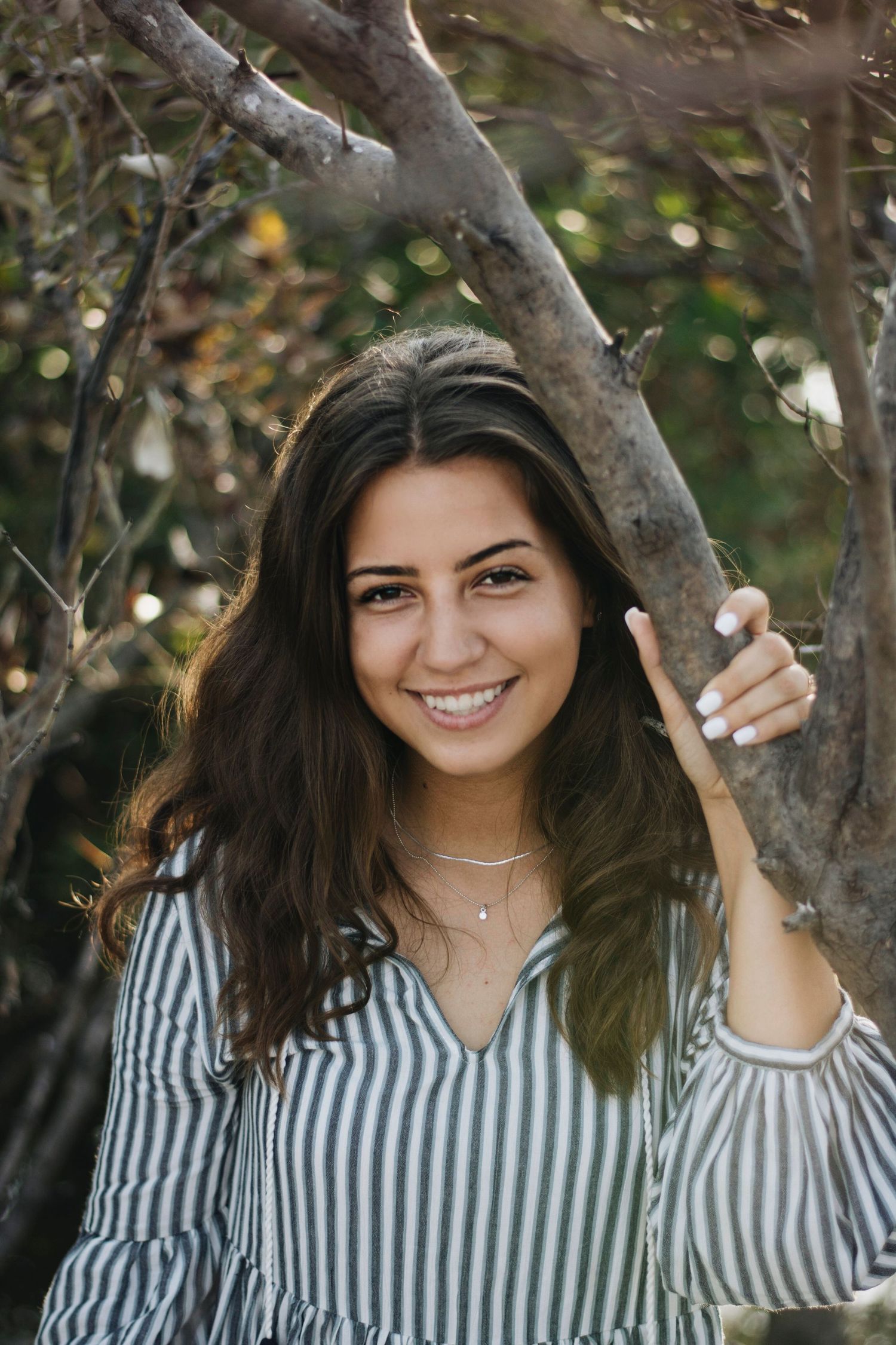Portrait of young woman in striped blouse posing by tree branch outdoors.