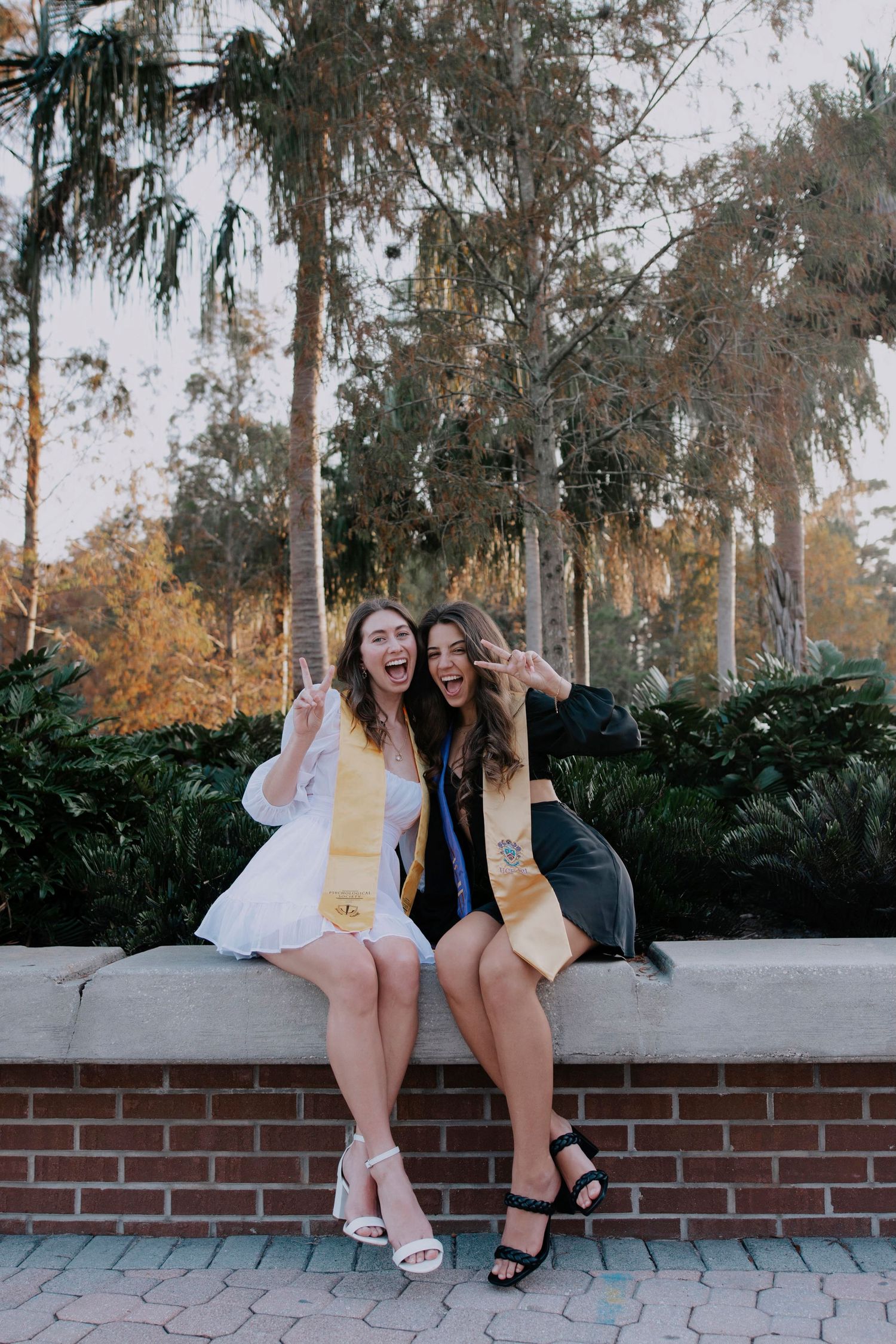 Two friends sitting on a brick wall outdoors sharing laughs in contrasting outfits.