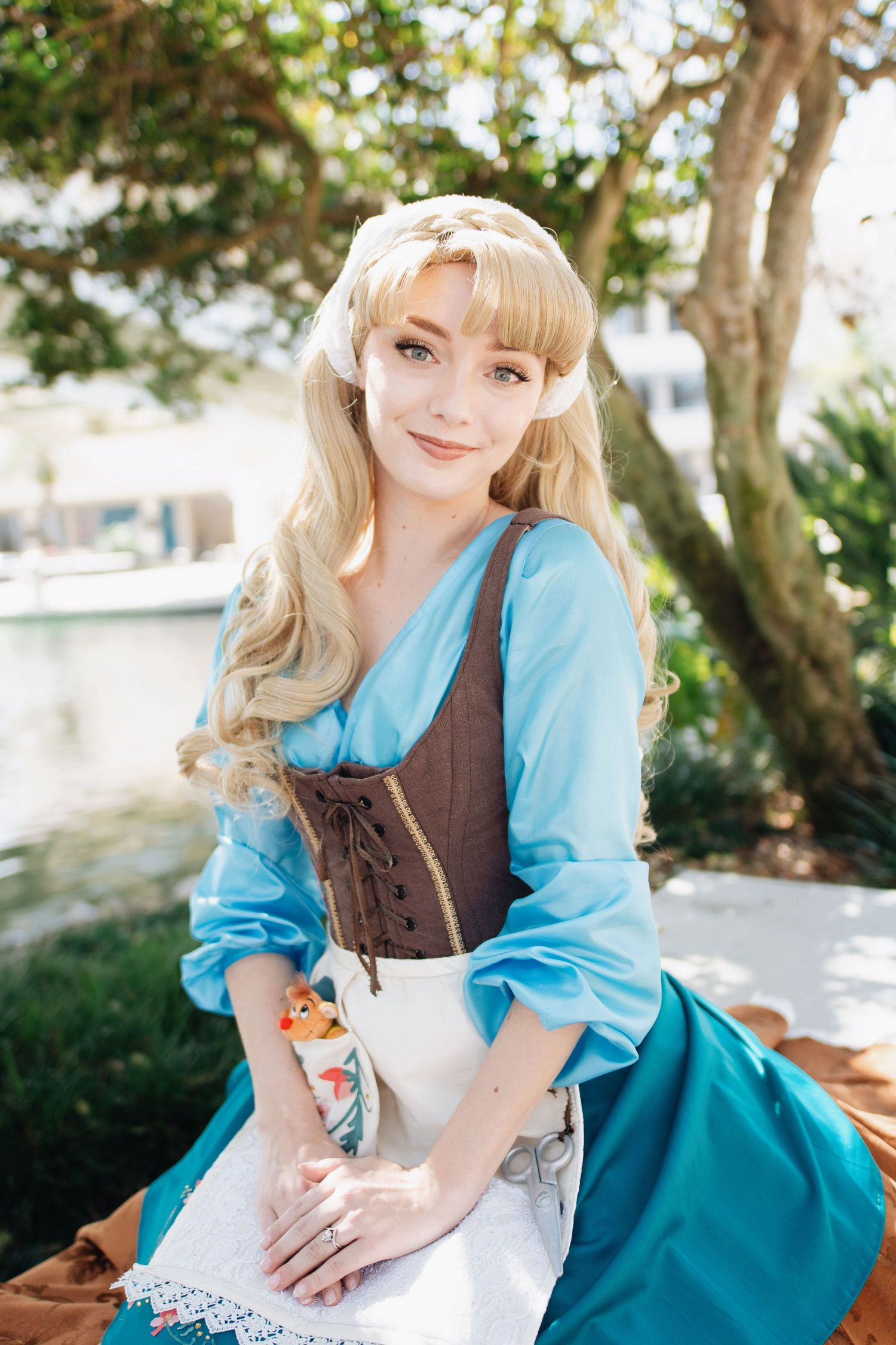 A figure in period costume with blue dress and brown corset poses in a sunny garden setting.