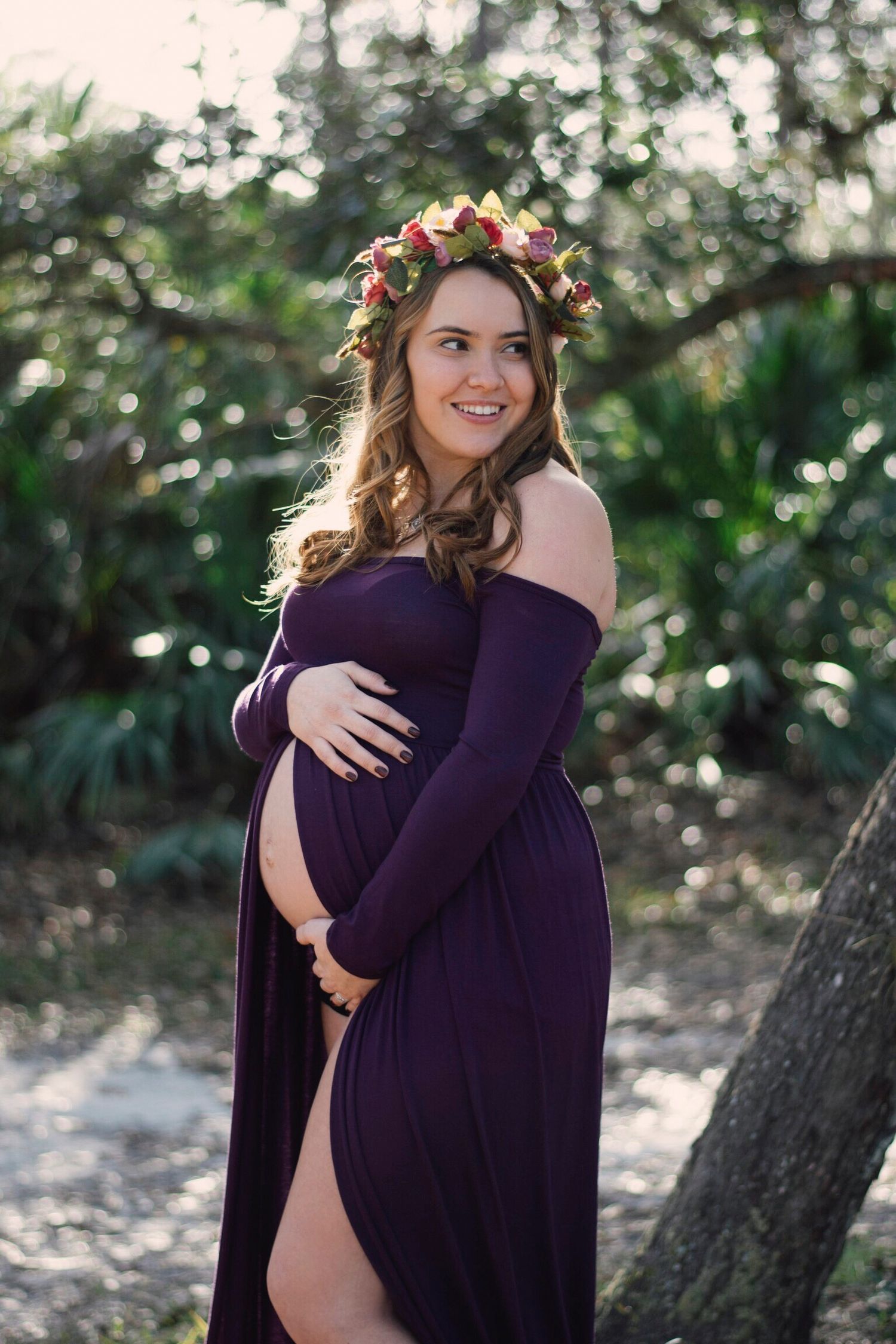 A pregnant woman in a purple dress poses in nature during a maternity photoshoot with sunlit trees in background.