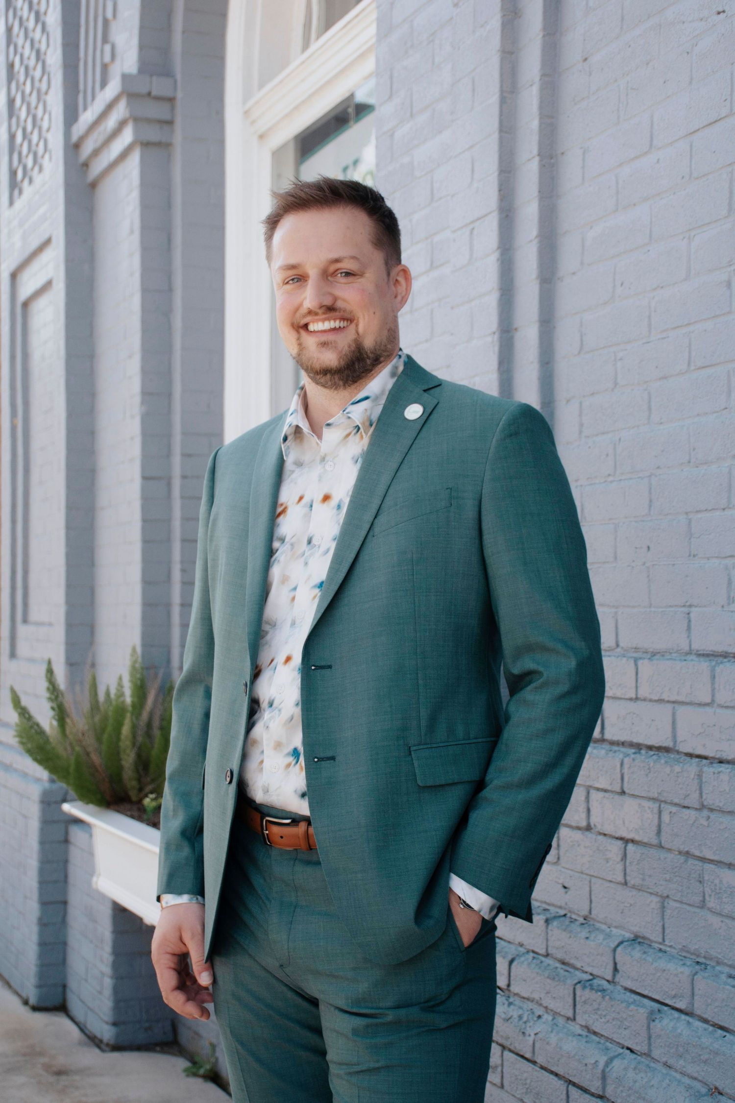 Full length business portraits in green suit against white brick building exterior.