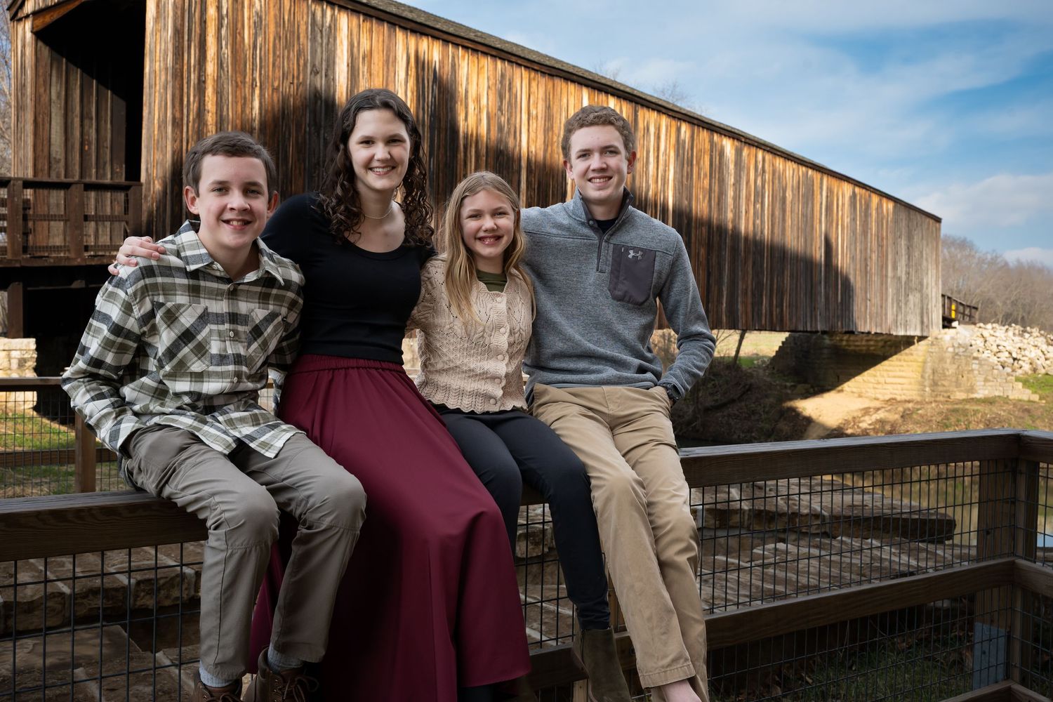 Family group poses outdoors in front of a rustic wooden barn structure on a sunny day.