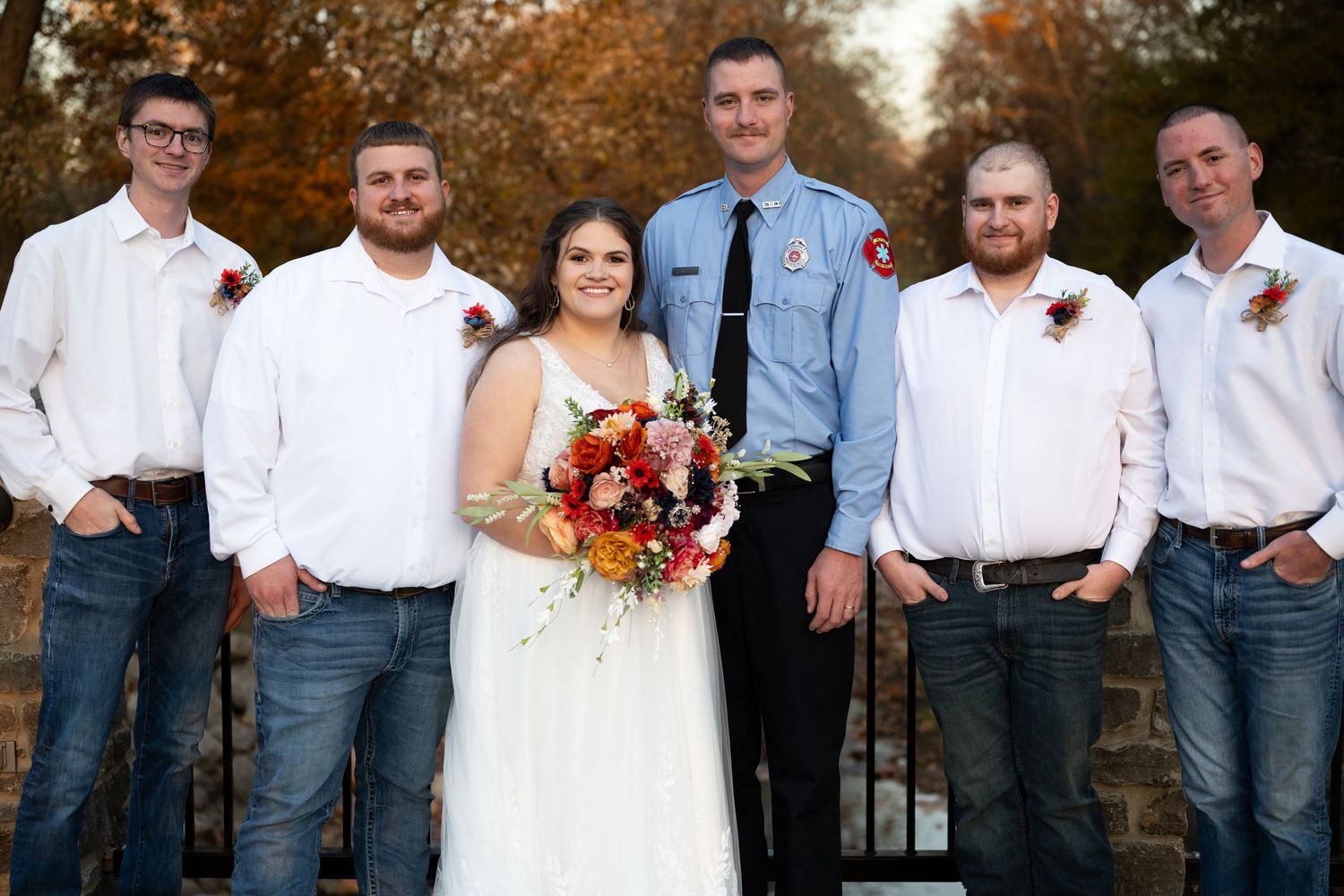 Wedding party members in casual attire pose together outdoors during autumn.