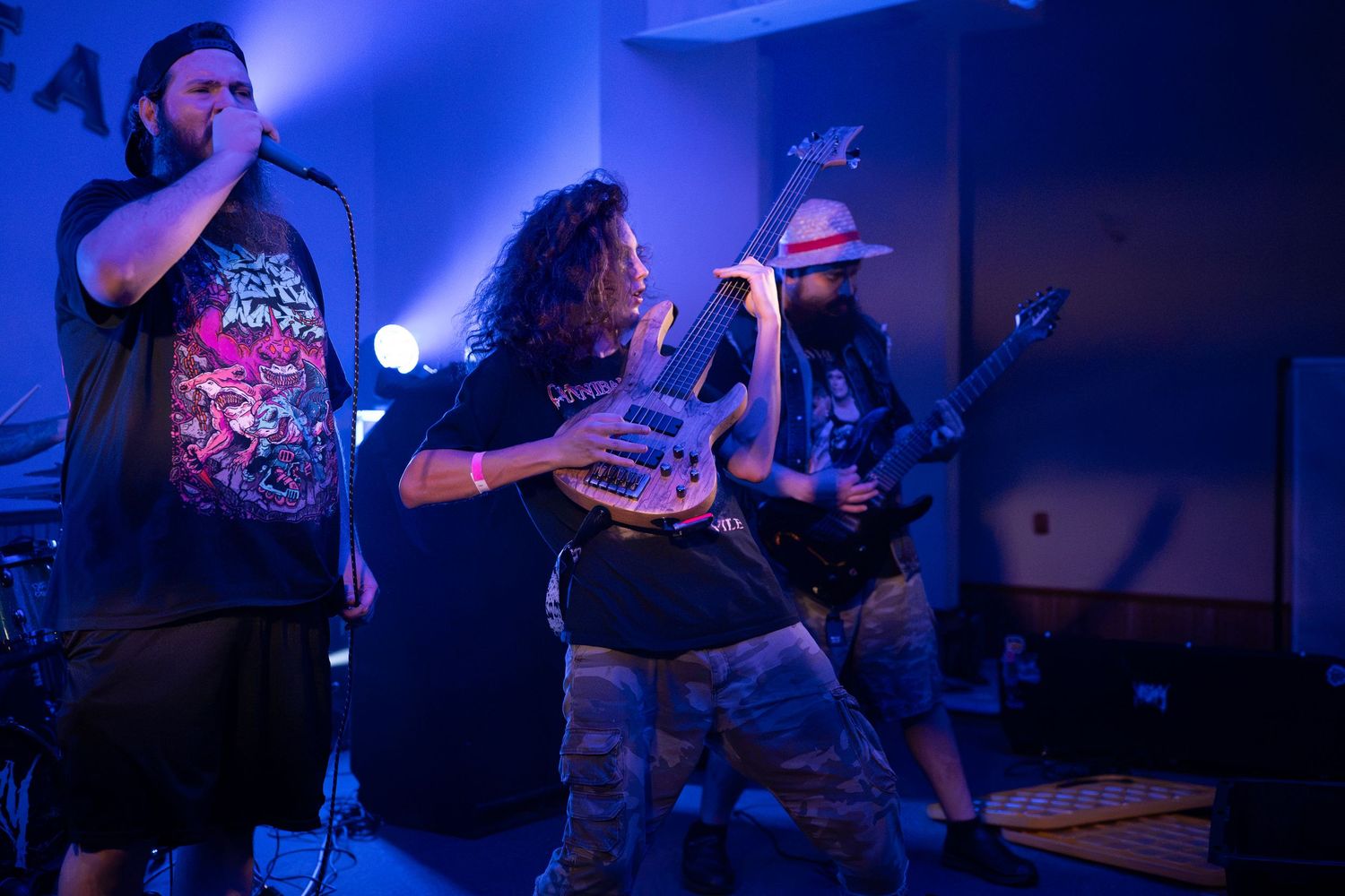 Three metal band members performing live on stage under blue and purple lights in an indoor venue.