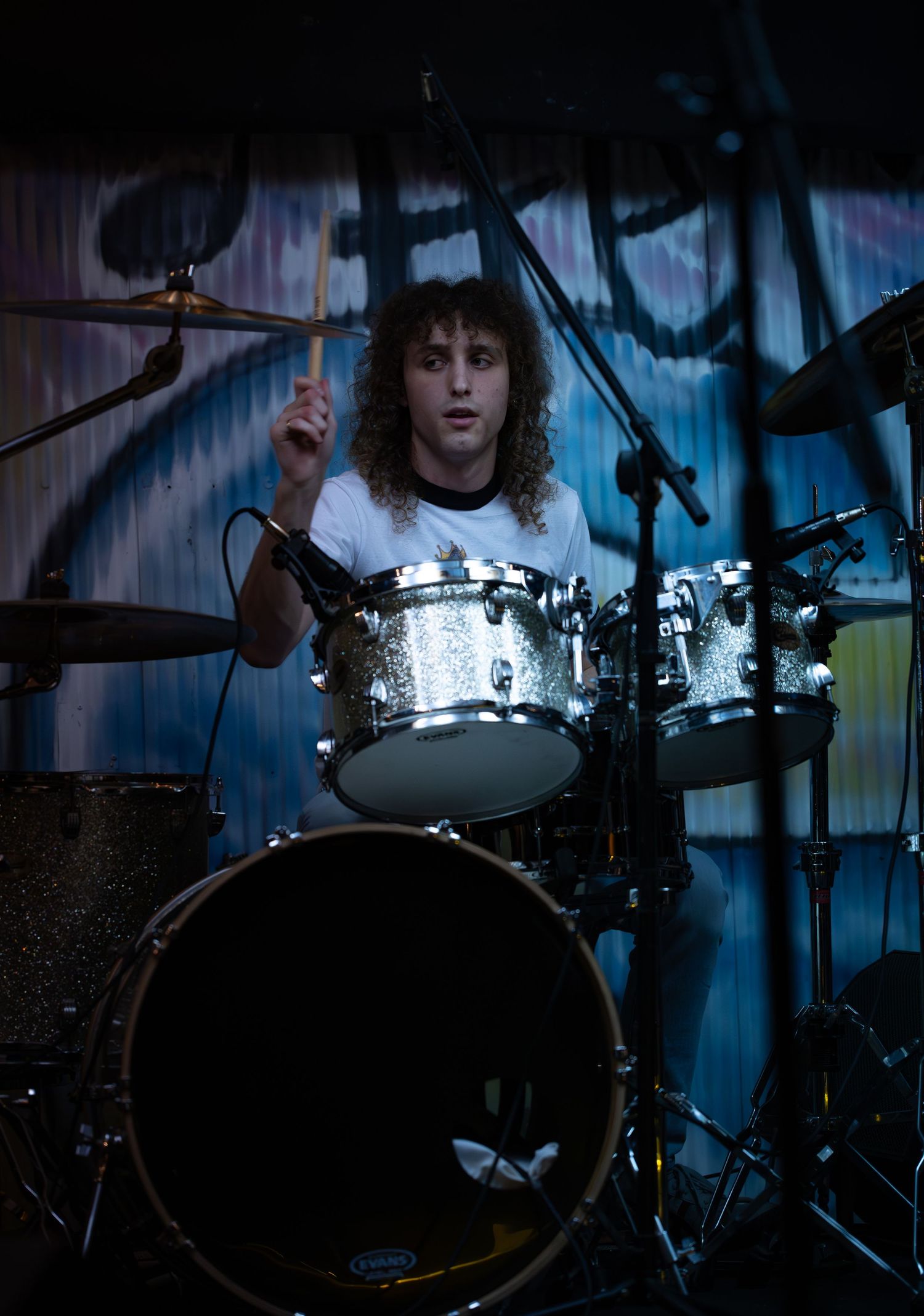 Drummer performs behind a sparkly silver drum kit on stage with blue lighting and dramatic shadows.