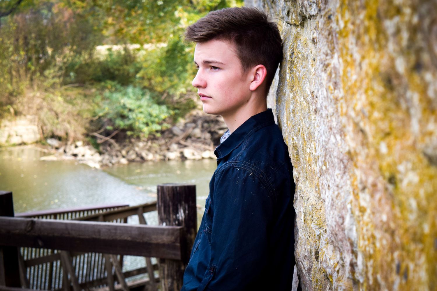 A person in a navy jacket leans against a tree trunk near a wooden deck overlooking water in soft natural lighting.
