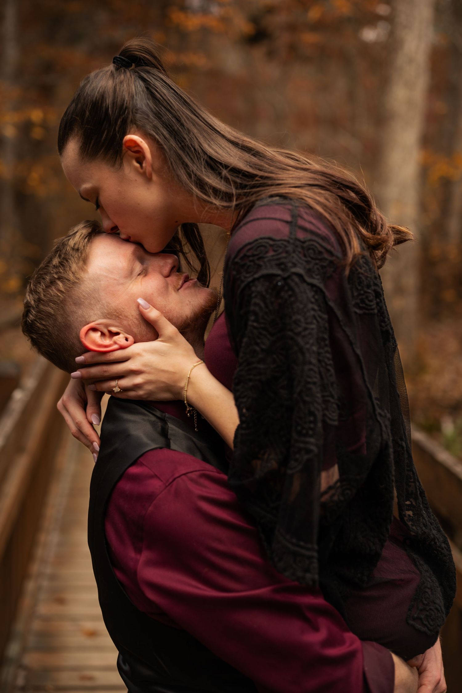A romantic couple sharing an intimate embrace in a warm burgundy and black outfit against an autumn forest backdrop.