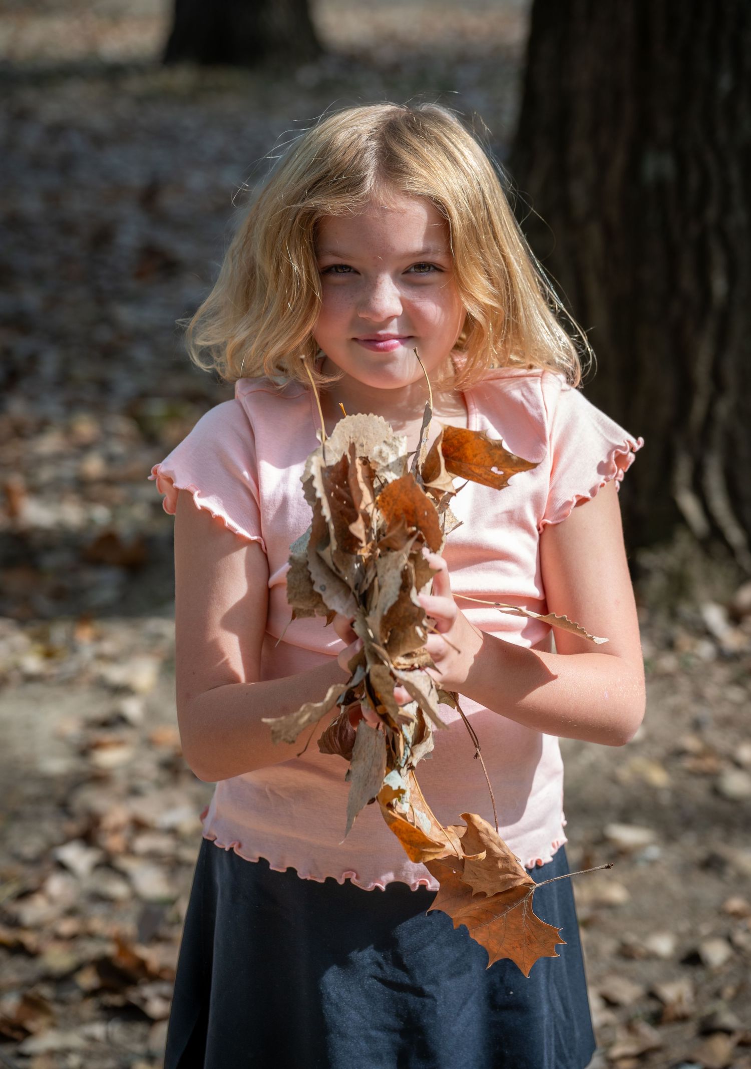 A person in a pink shirt and jeans holds dried autumn leaves while playing outdoors in a sequence of four photos.
