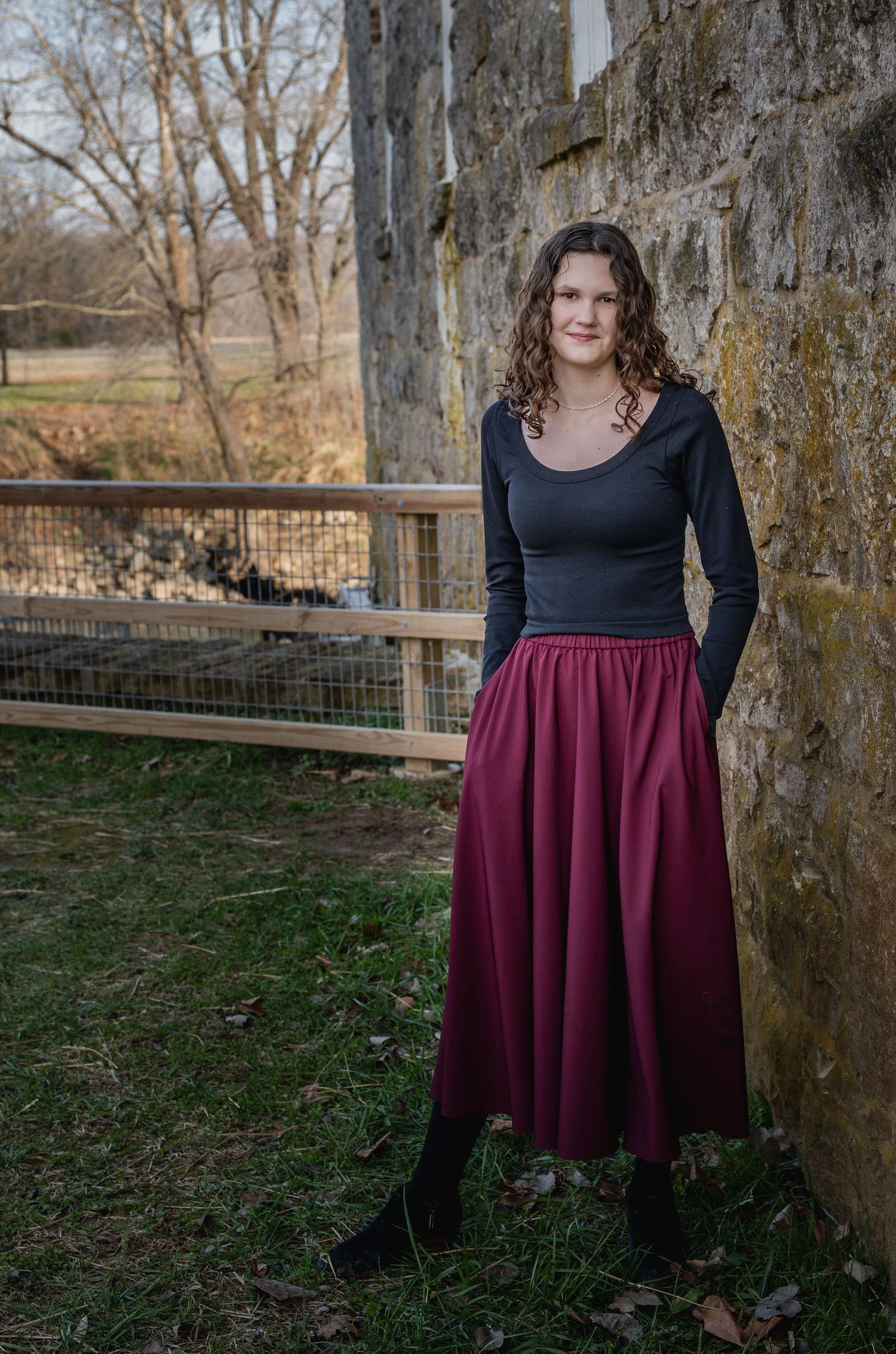 Person in black top and burgundy skirt standing near rustic wooden posts outdoors.