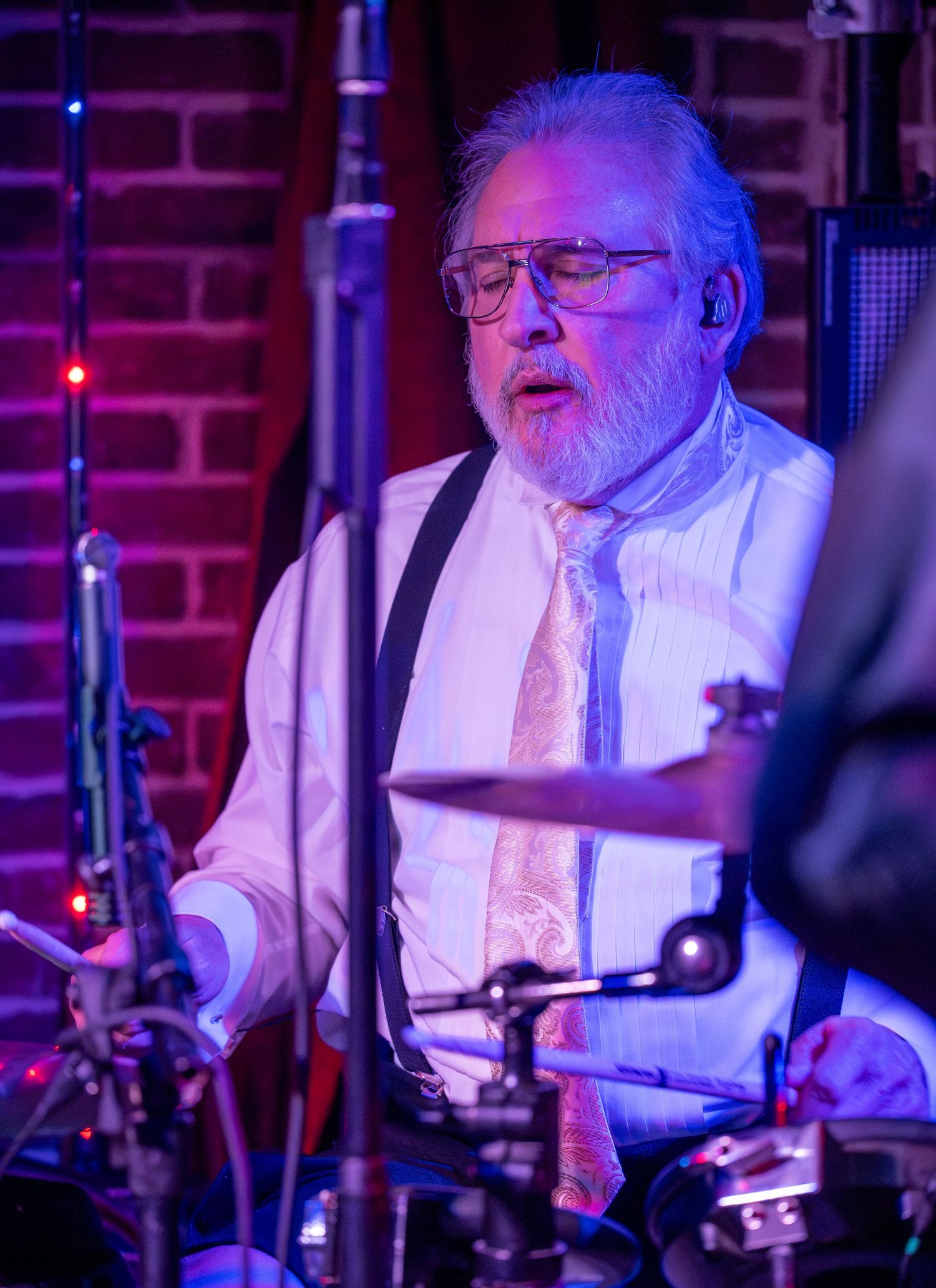 Drummer in white shirt and suspenders performs at a brick-walled venue with purple stage lighting.