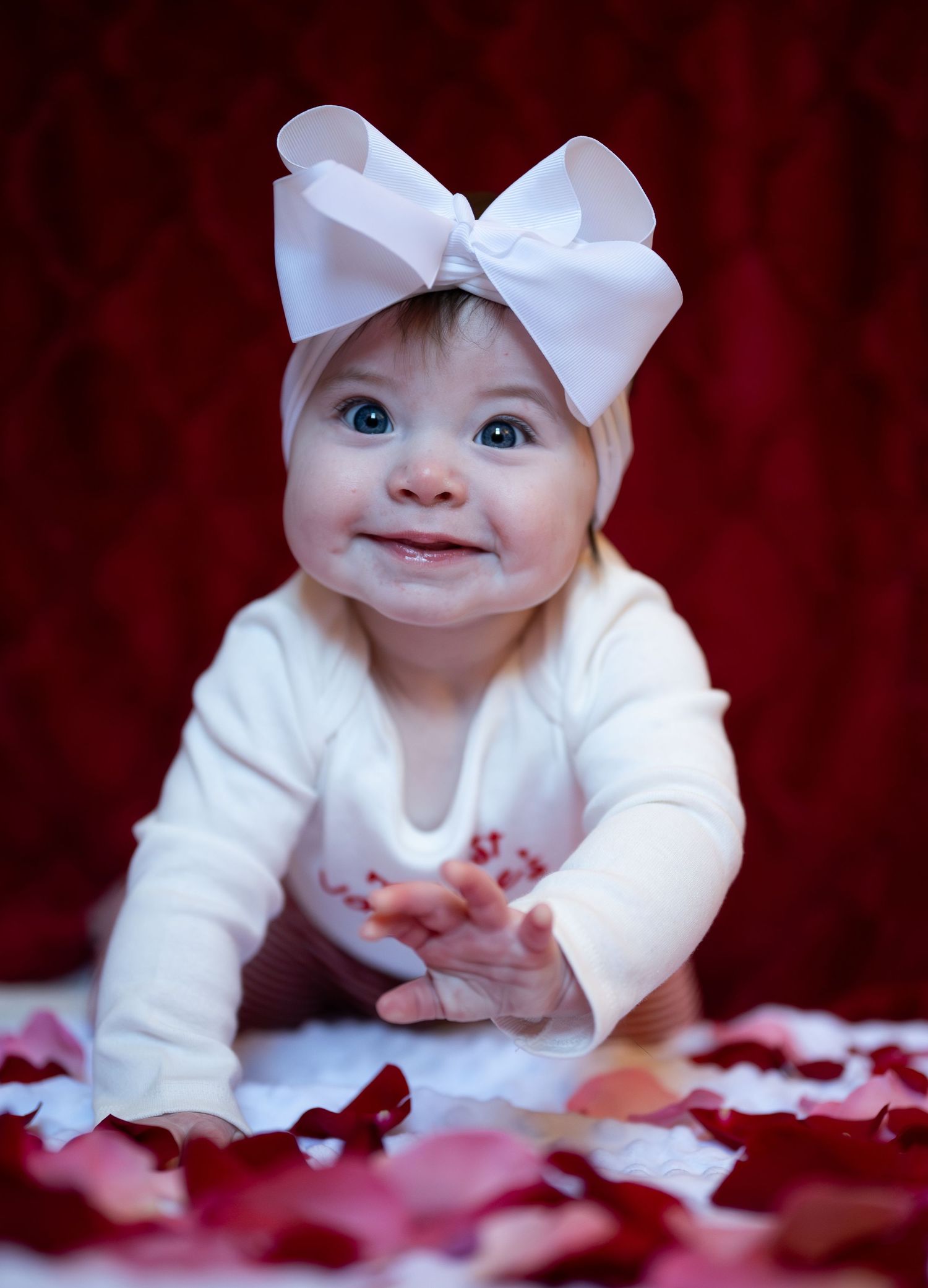 Baby in white outfit with large white bow headband smiles while lying on red backdrop with scattered rose petals.