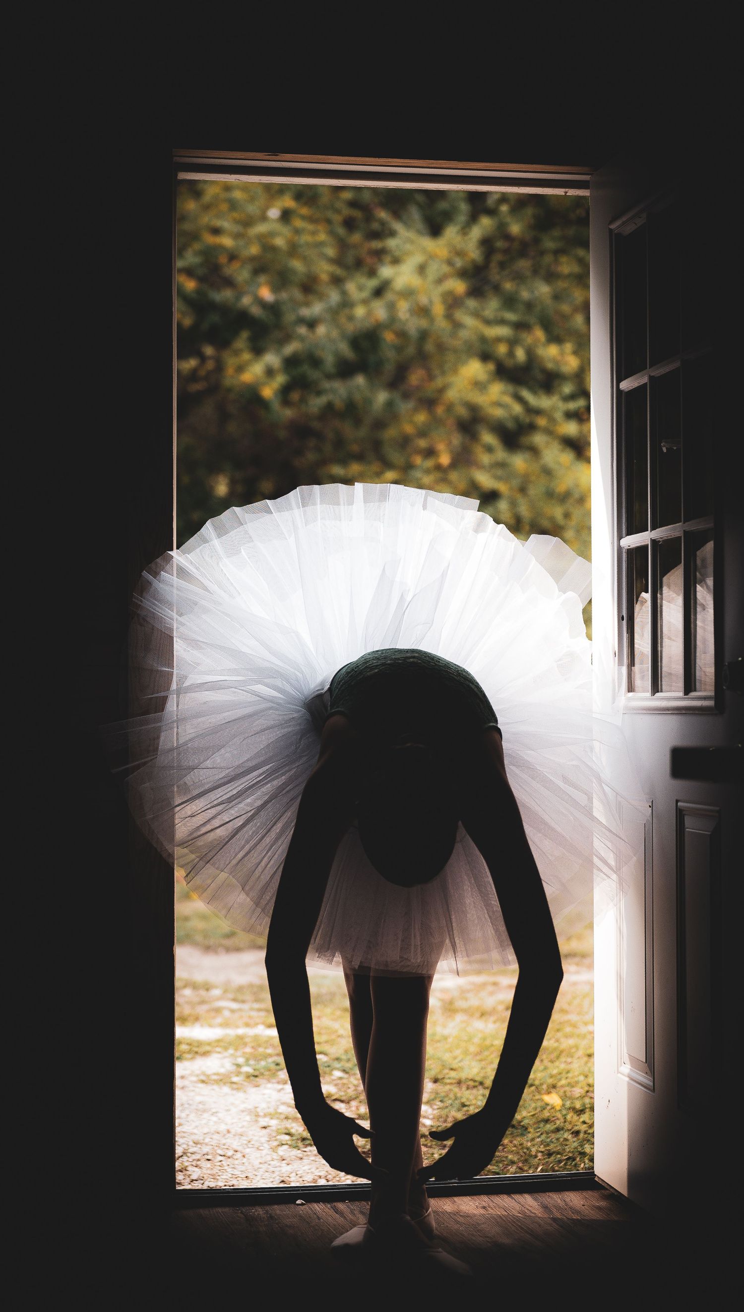 A silhouetted figure in a tutu bends forward in a doorway creating a dramatic backlit ballet pose.