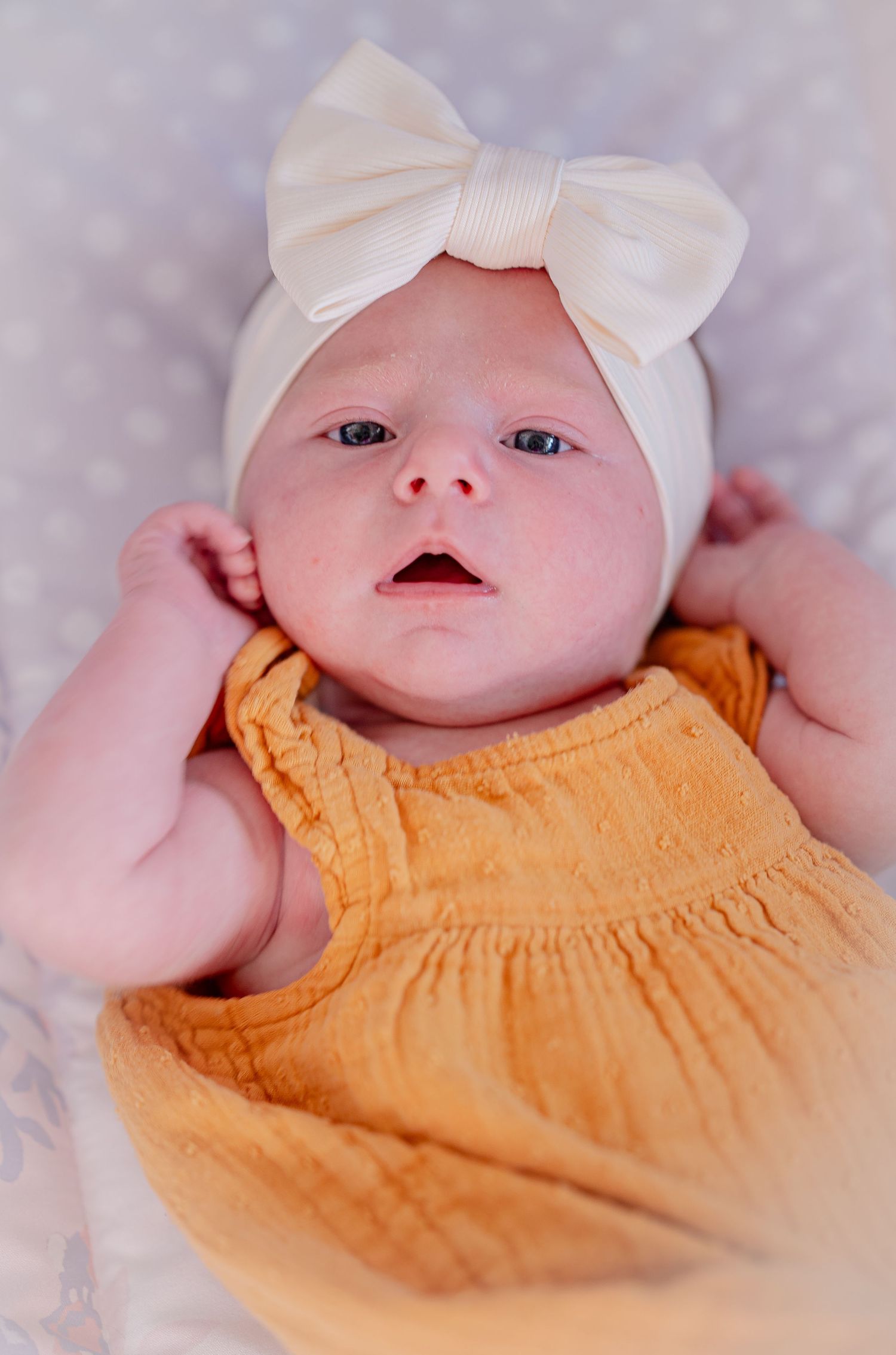 A baby wearing a white bow headband and orange dress smiles while lying on soft bedding.