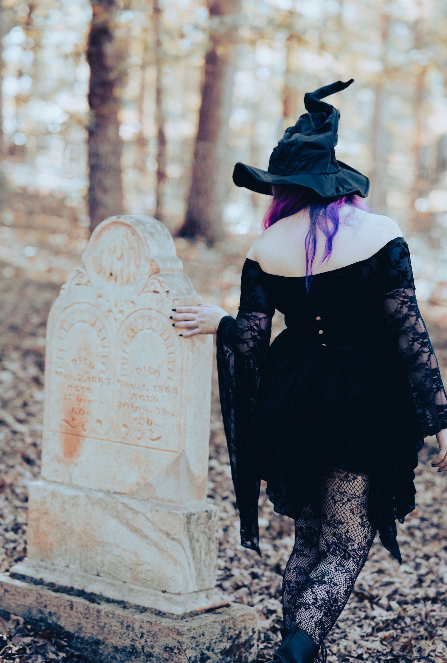 A witch in black lace dress and pointed hat stands beside an old weathered gravestone in an autumn forest.