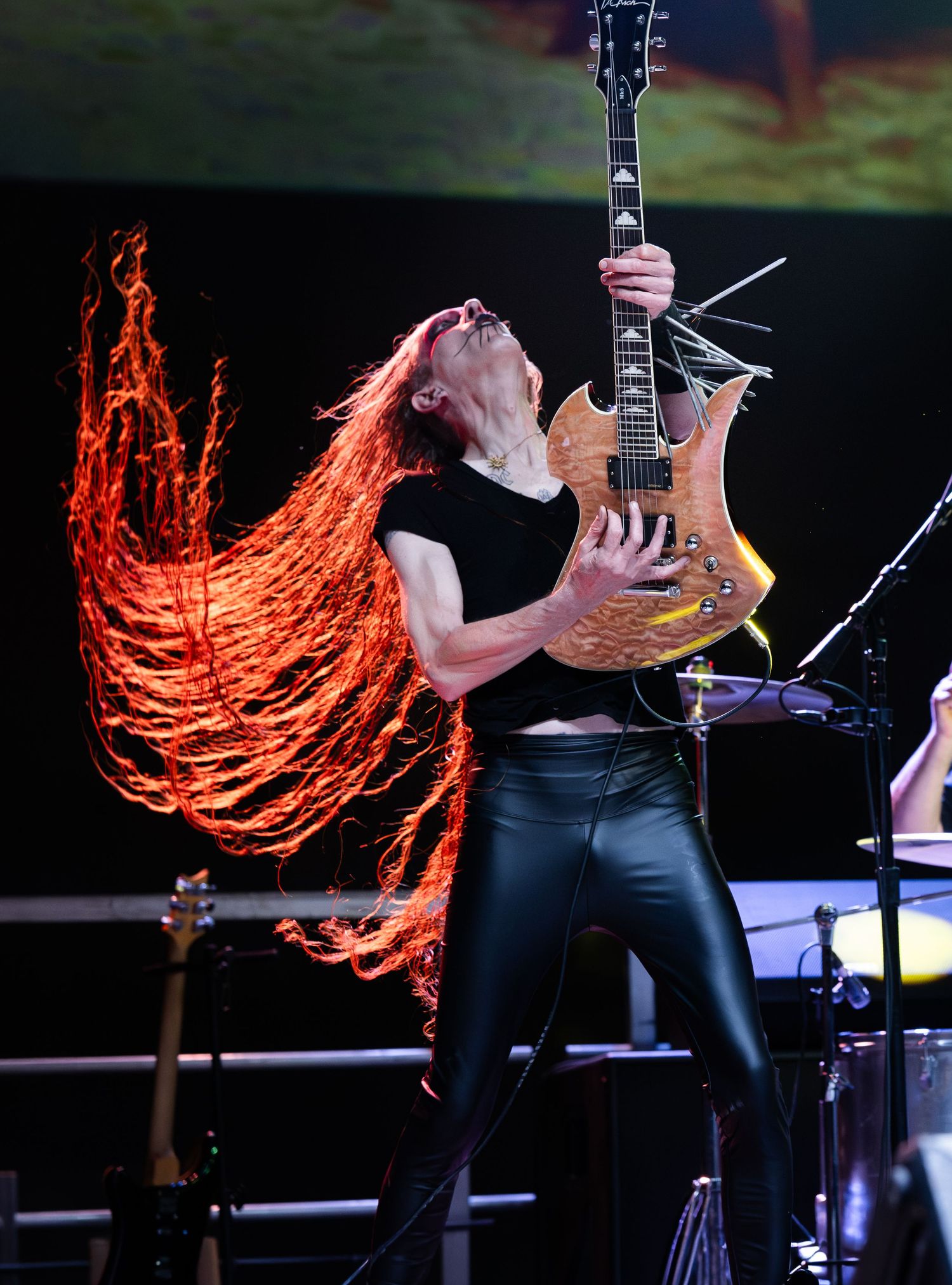 Female guitarist with flowing red hair plays electric guitar on stage in black outfit during energetic live performance.