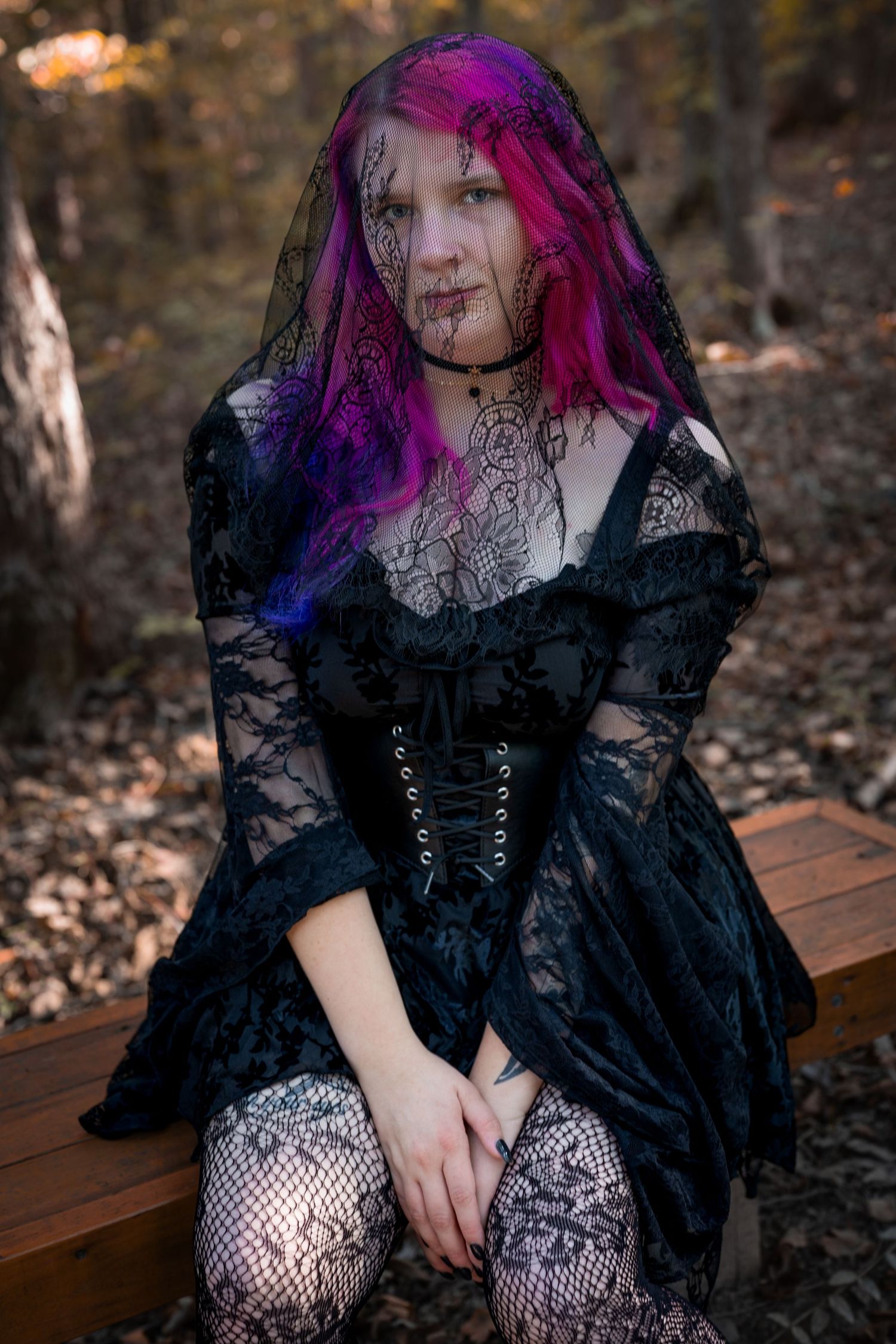 Gothic style portrait featuring black lace dress, patterned stockings, and vibrant purple hair in a moody forest setting.