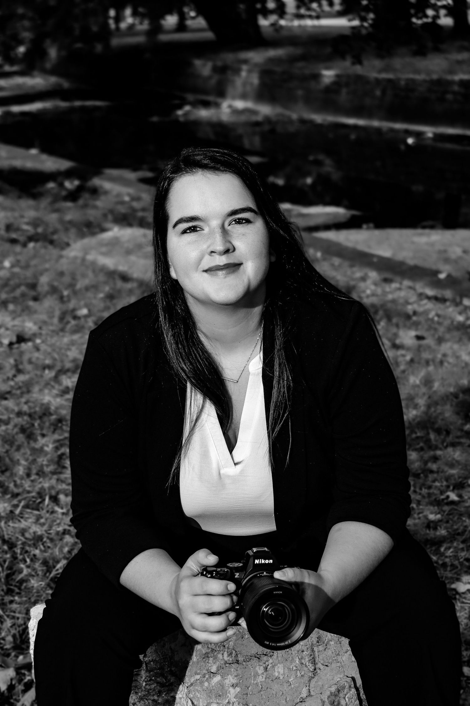 Black and white portrait of photographer sitting outdoors holding a camera while smiling at the camera.