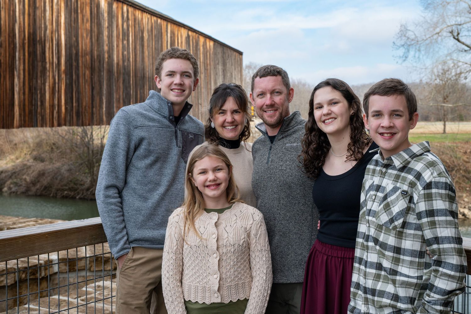 A group of people pose together outdoors in casual winter attire near wooden structures and natural scenery.