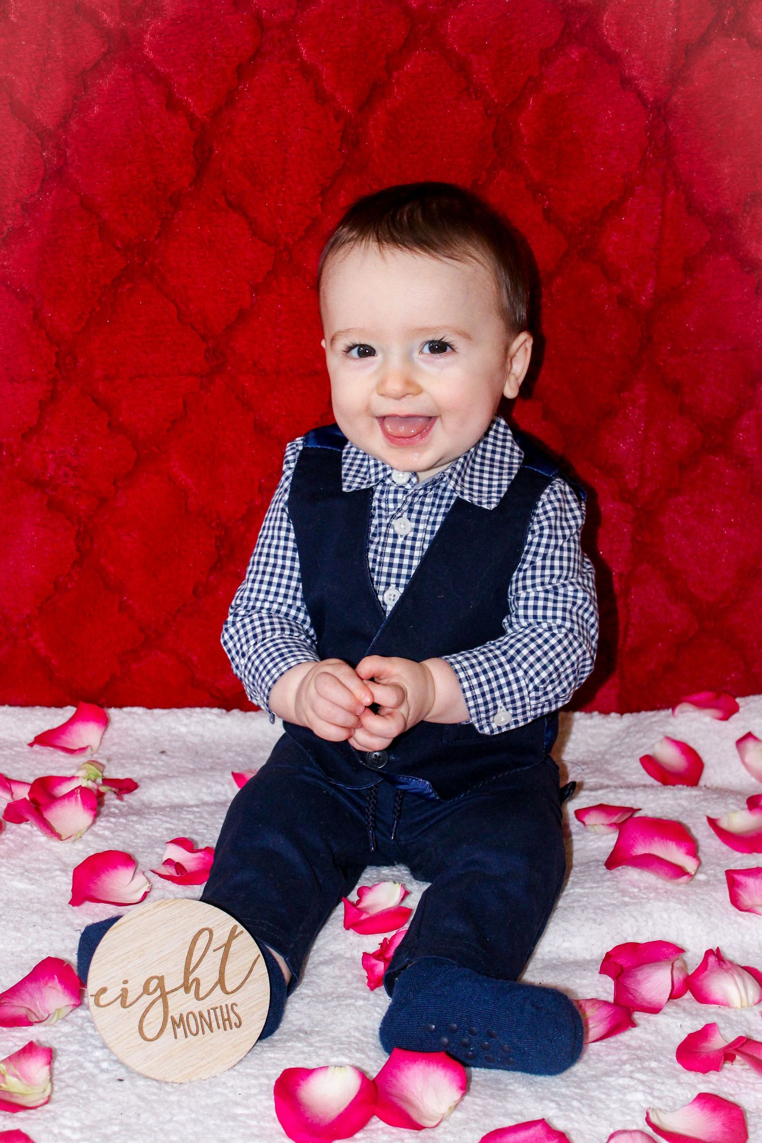 Baby in navy vest and gingham shirt sits on white blanket with pink rose petals against red backdrop.