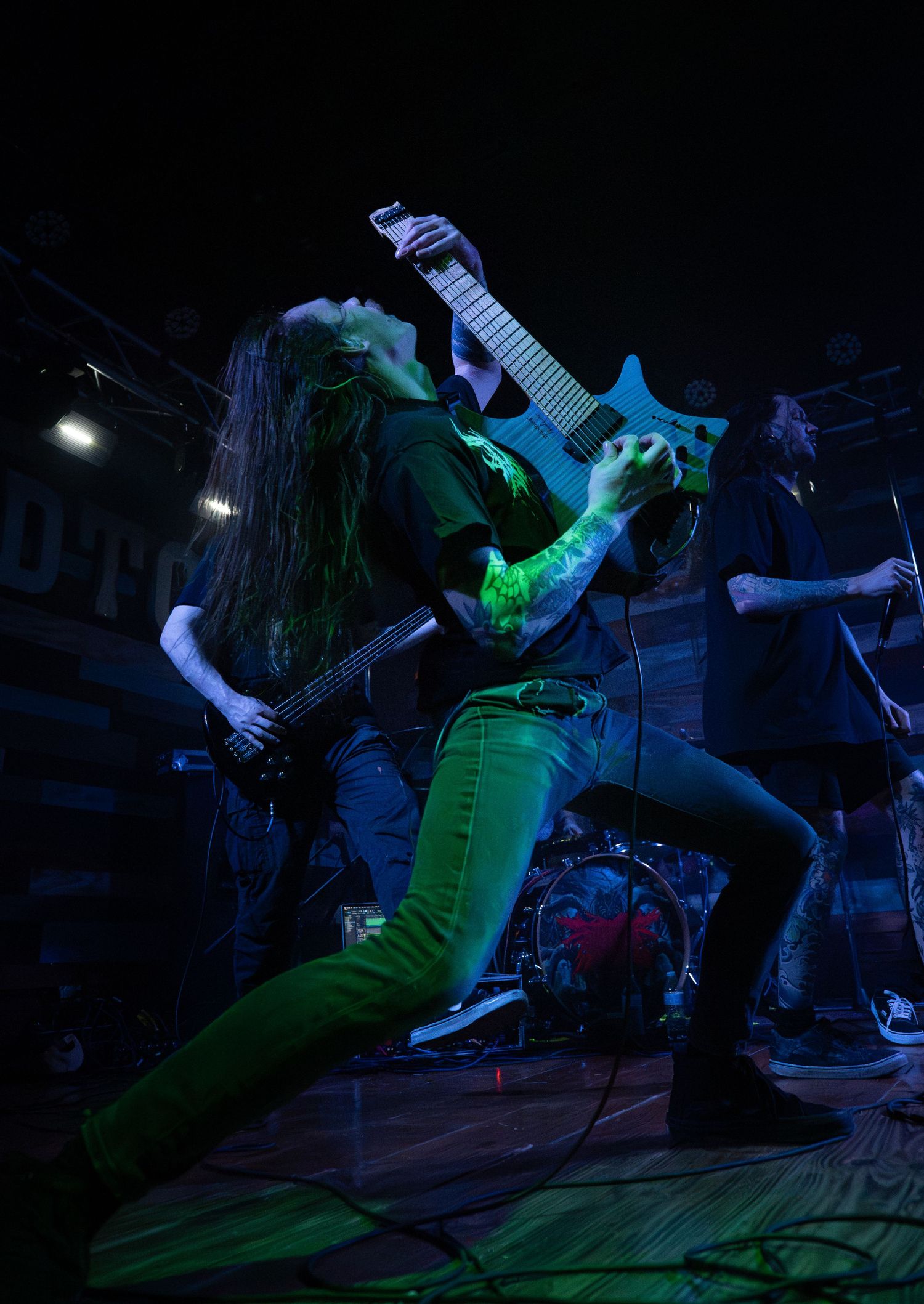 Metal guitarist performs on stage under green lights, playing electric guitar with long hair and green jeans.