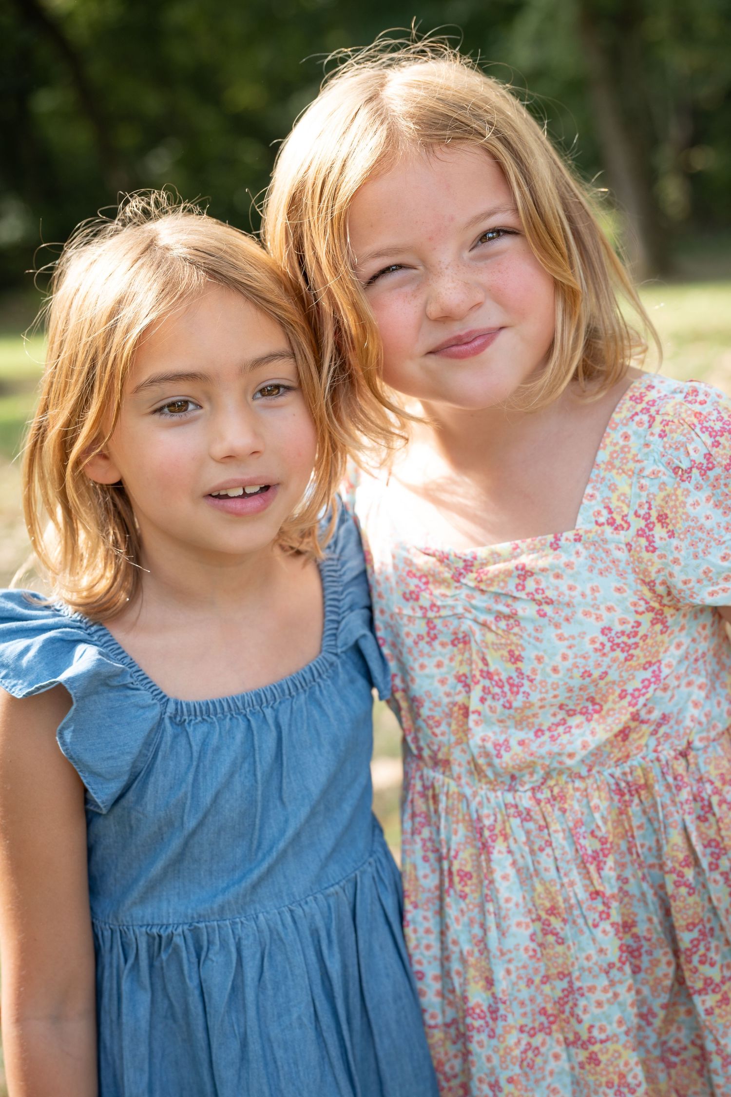 Two young children wearing summer dresses smile together outdoors on a sunny day.