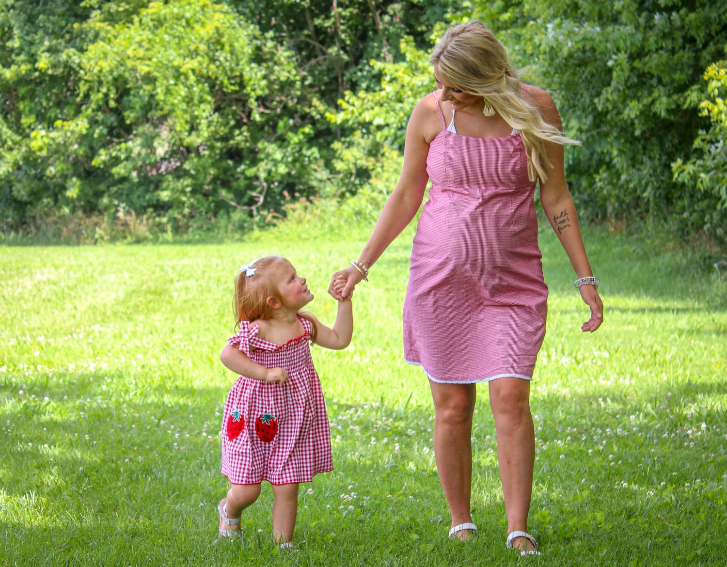 A parent and child in pink outfits walk together on a lush green lawn during a sunny summer day.