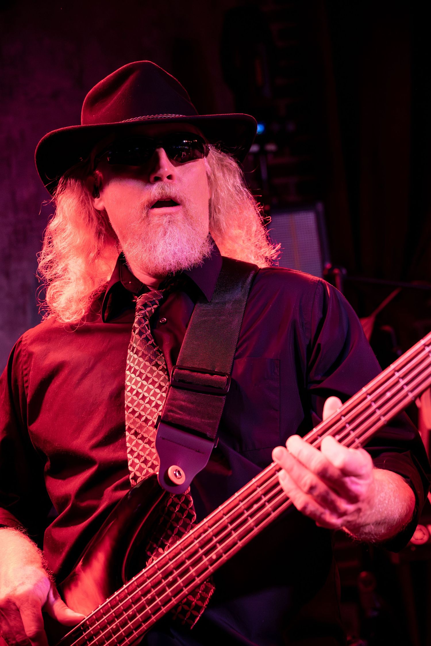 A bass guitarist in a black hat performs on stage under dramatic red lighting during a concert performance.
