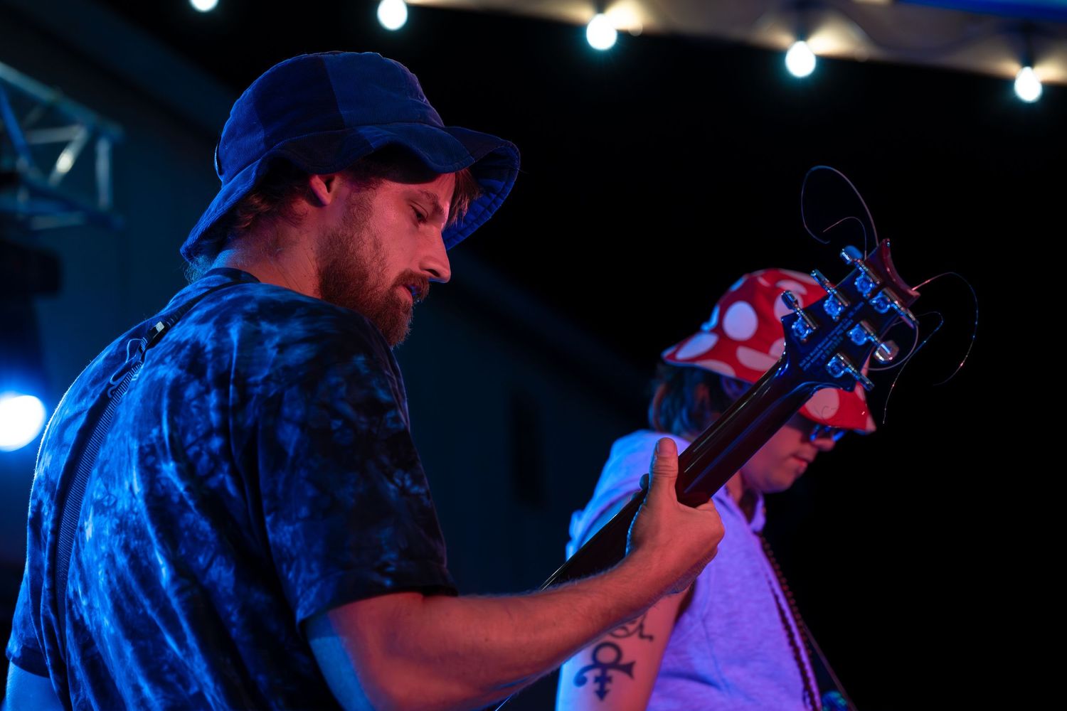 Musician in blue cap and plaid shirt plays guitar on stage under bright spotlights during performance.