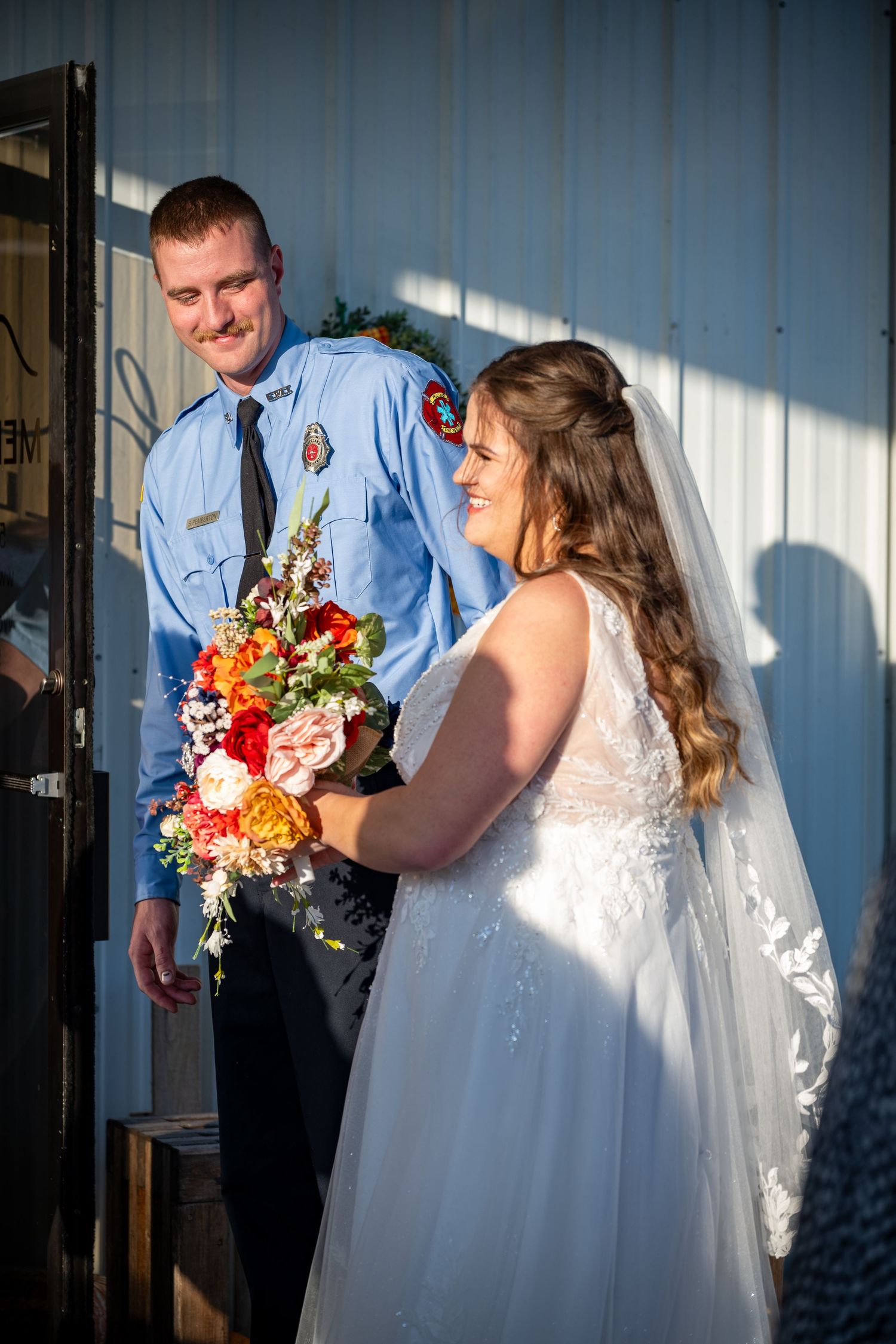 Wedding couple in white dress and blue uniform stand in dramatic lighting.