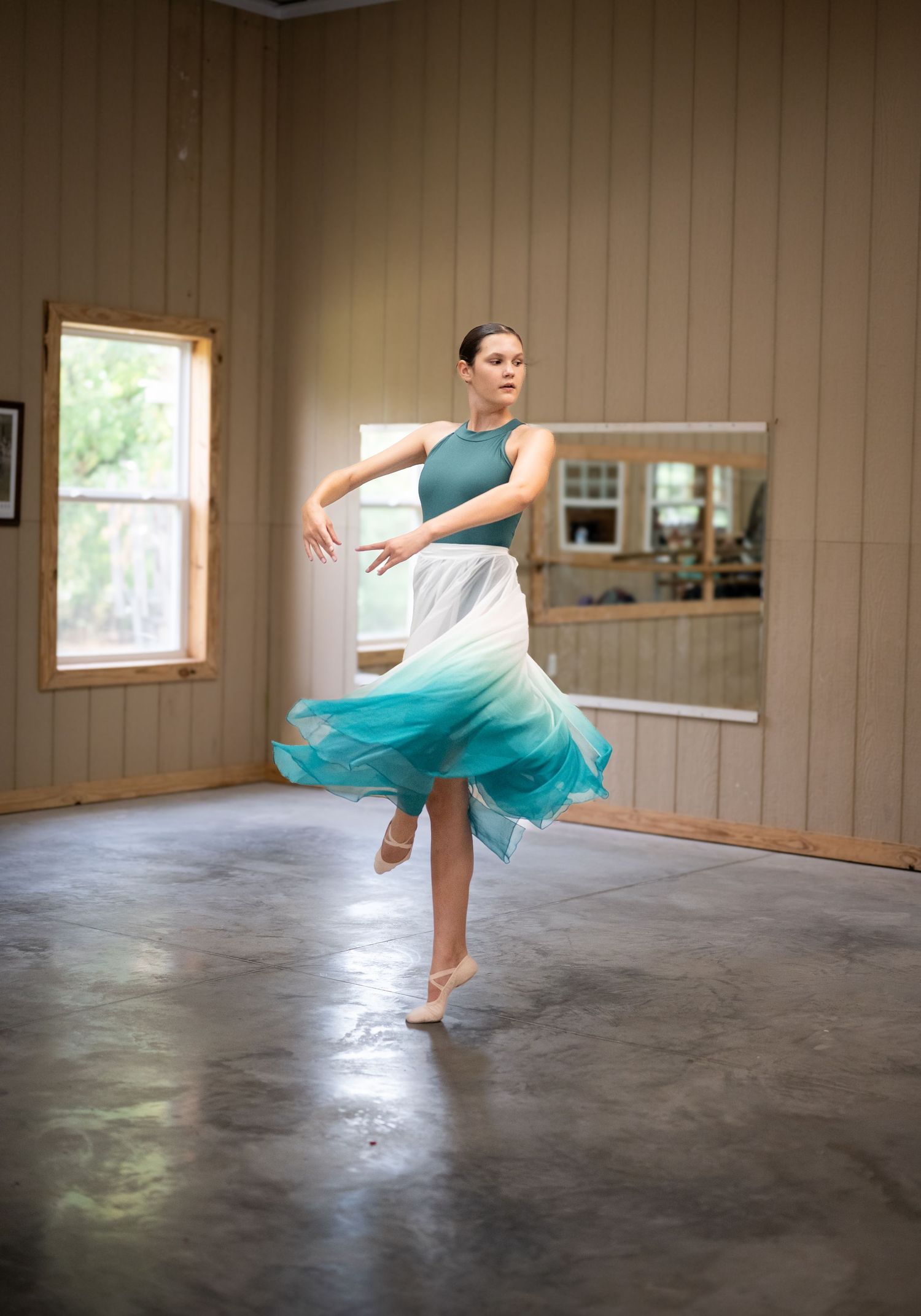A dancer in a flowing turquoise and white dress performs a graceful spin in a dance studio with wooden walls and natural light.
