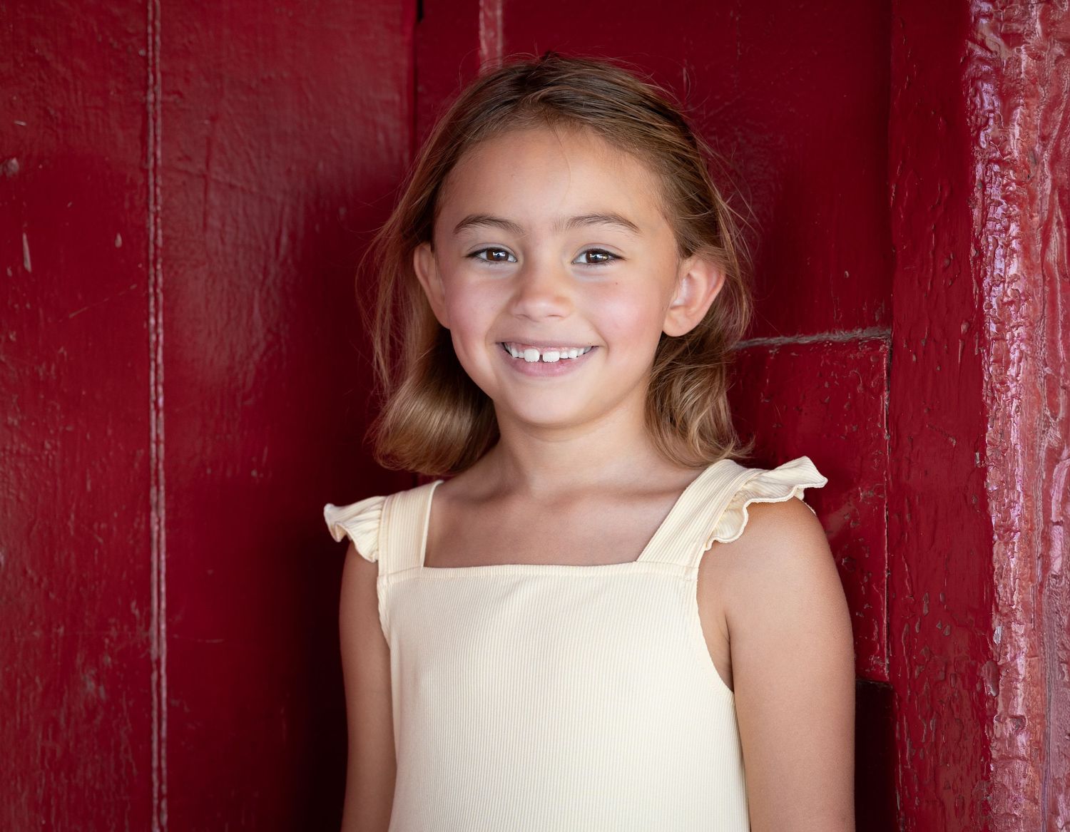 A child in a cream-colored dress poses against a deep red wooden wall with a bright, cheerful expression.