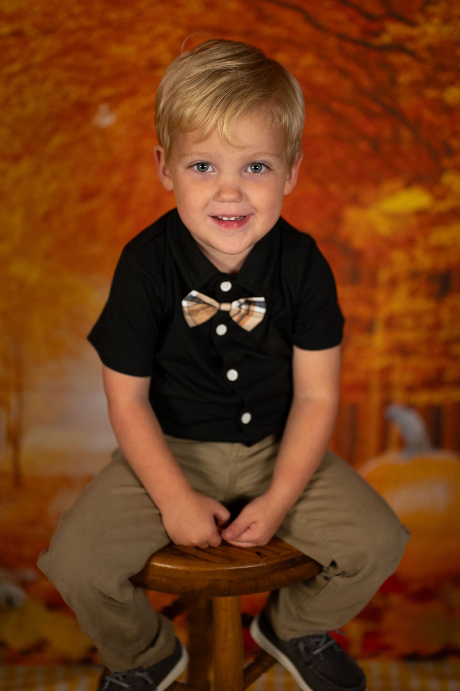 A child in black shirt and khaki pants sits on a wooden stool against an autumn-colored backdrop.
