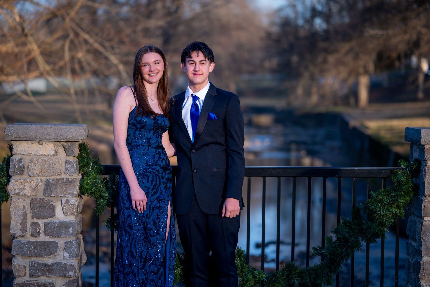A couple in formal attire poses for romantic outdoor photos near a stone wall and railing at sunset.