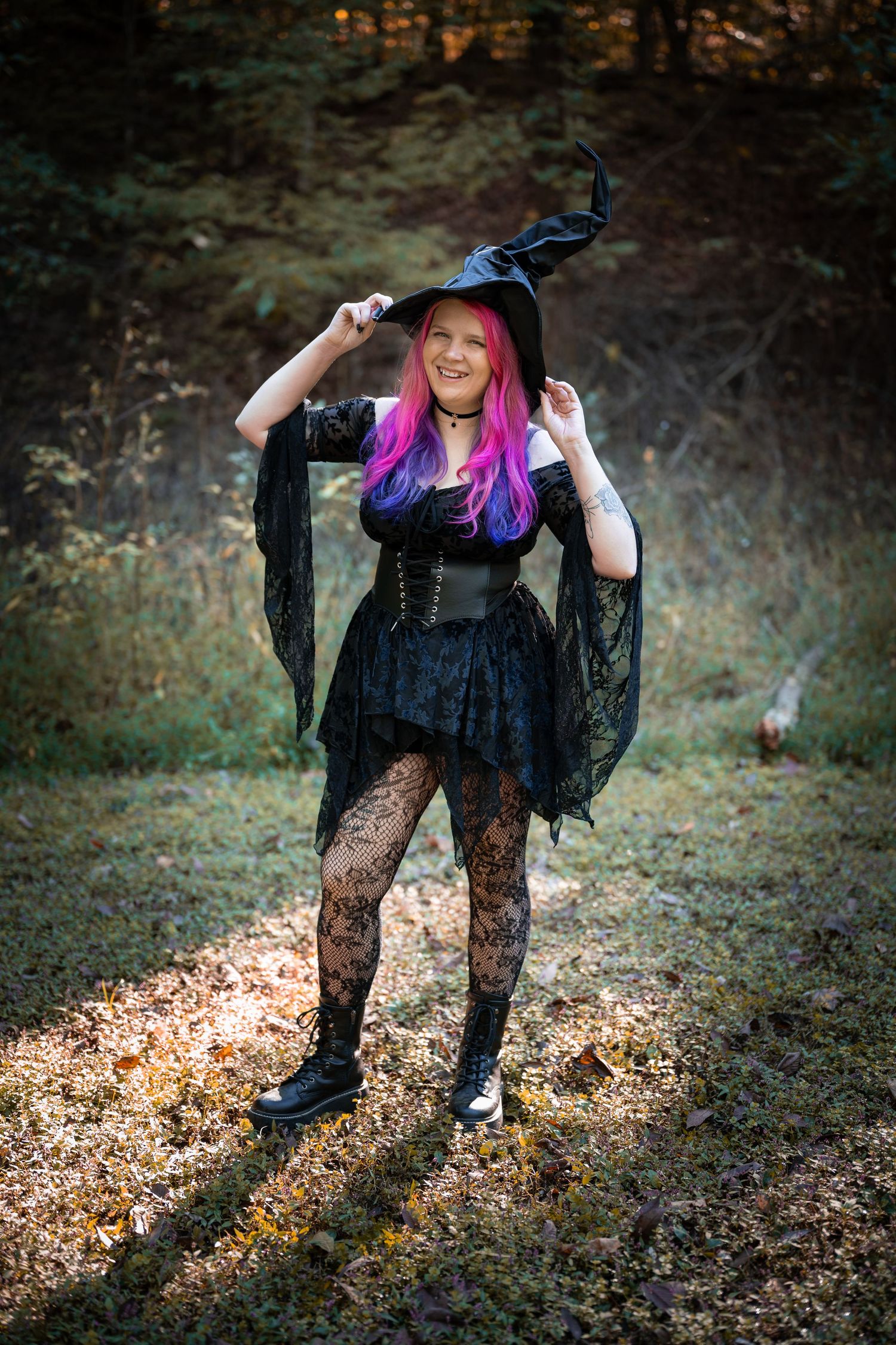 A person in a black witch costume with colorful hair poses playfully on autumn leaves in a forest setting.