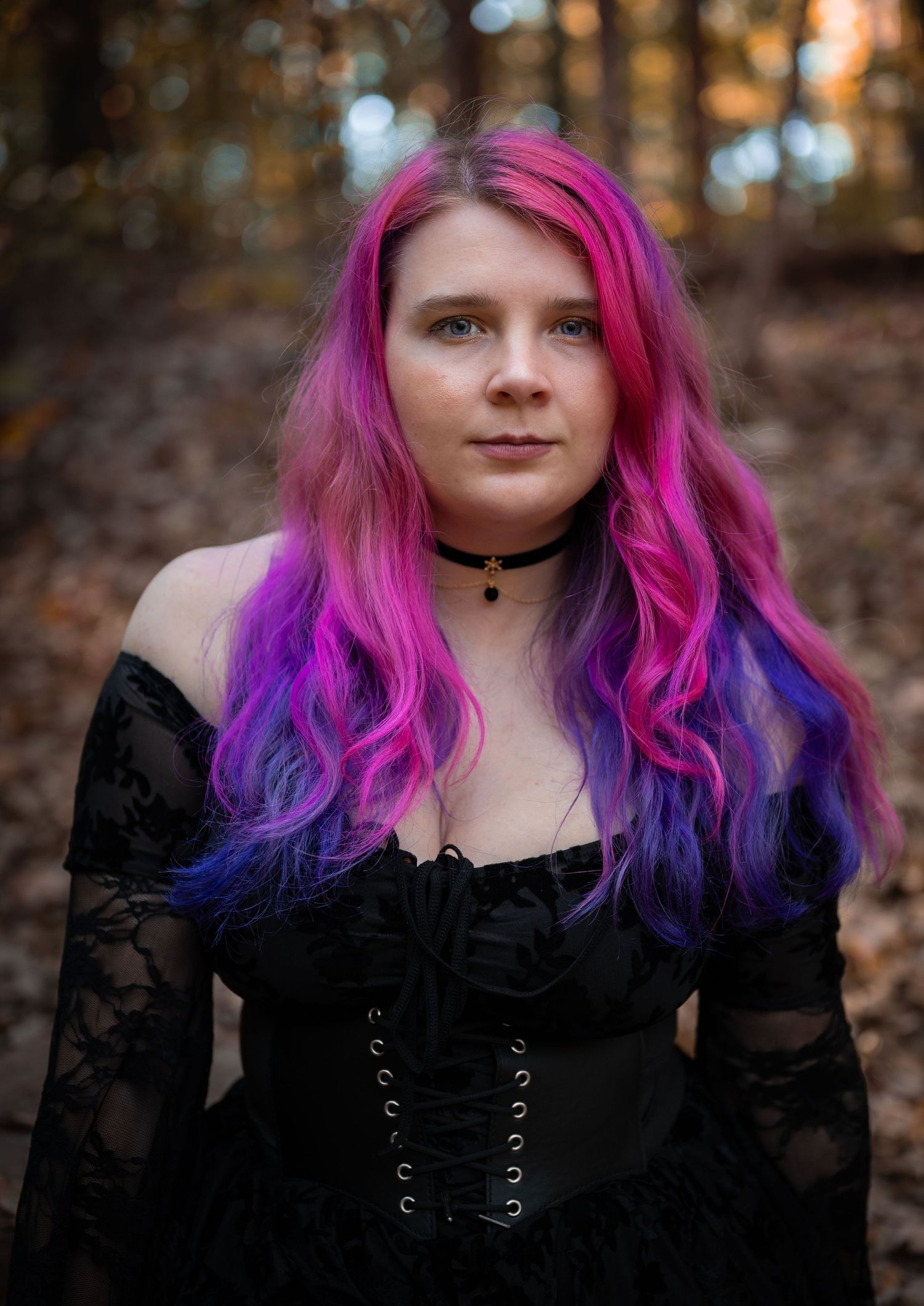 A person with vibrant pink and purple ombre hair wearing a black gothic-style dress poses in a wooded outdoor setting.
