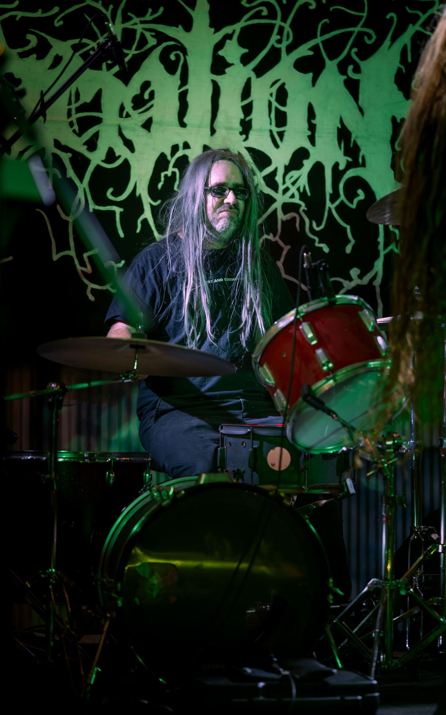 Drummer performs on stage with metal band setup illuminated by dramatic green lighting during live concert performance.
