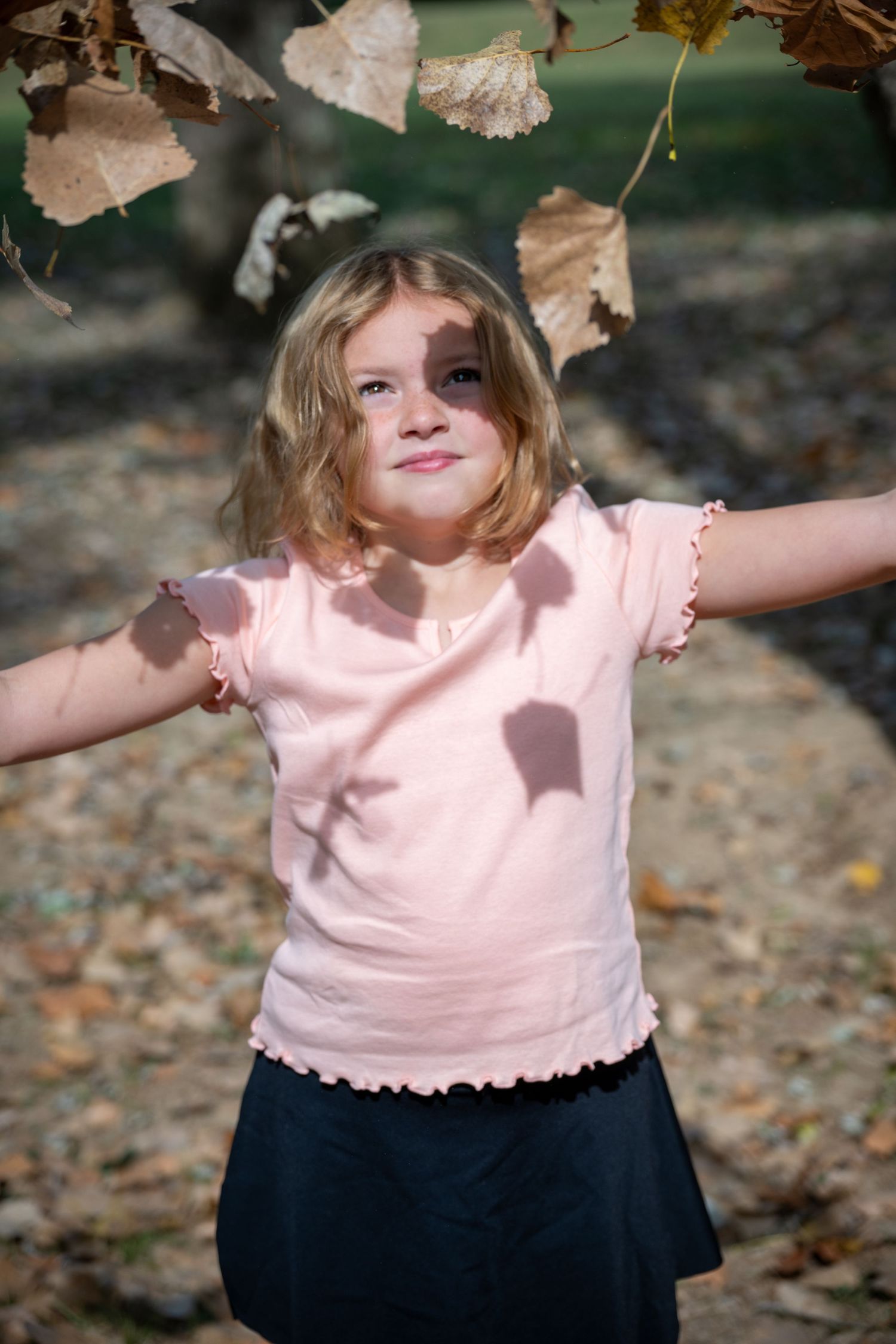 A child in a pink top and navy skirt plays joyfully among autumn leaves in an outdoor setting.