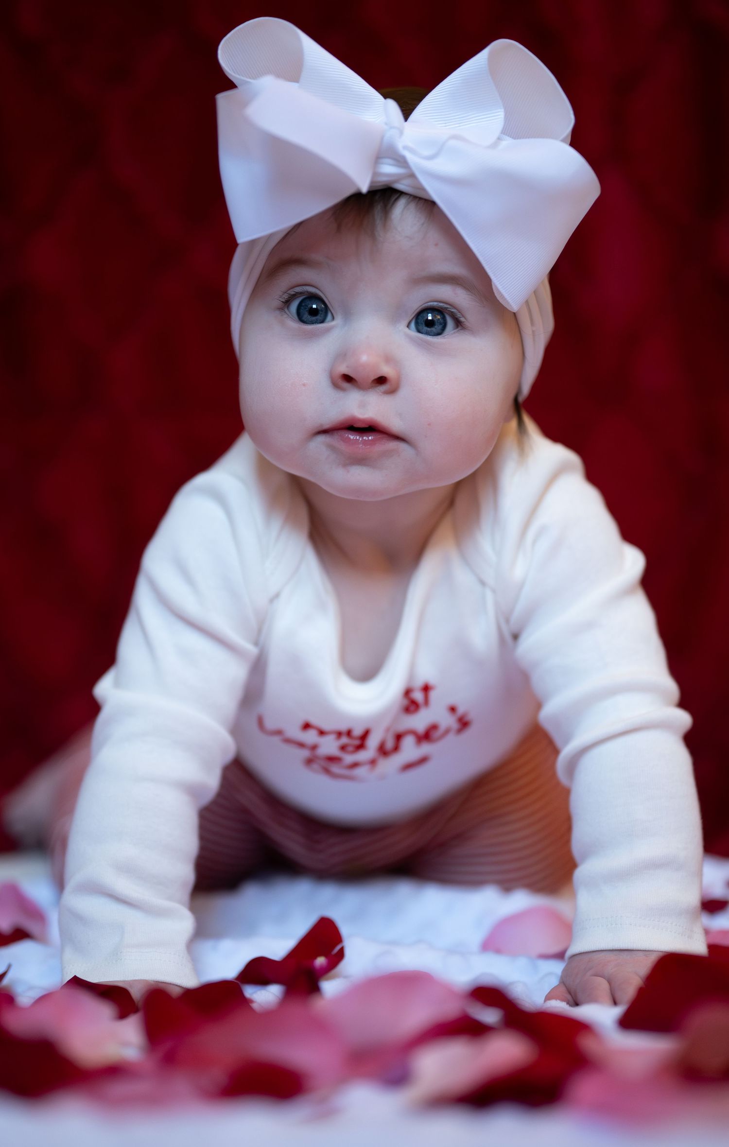 Baby in white outfit with bow headband lies on red background with scattered rose petals.