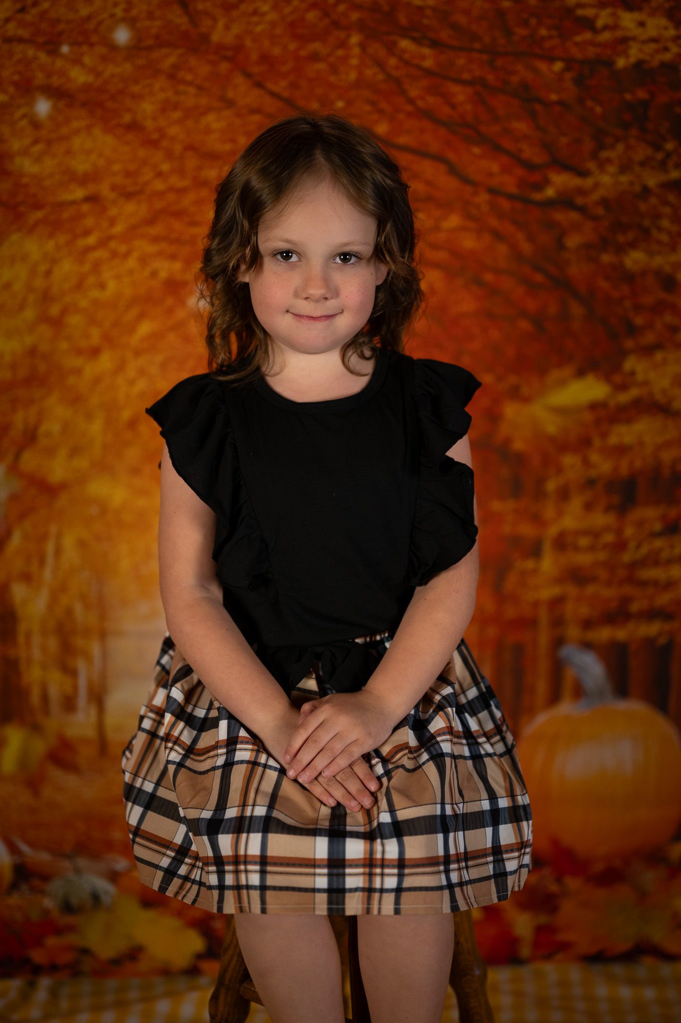A young child in a black ruffled top and plaid skirt poses against an autumn backdrop with pumpkins.
