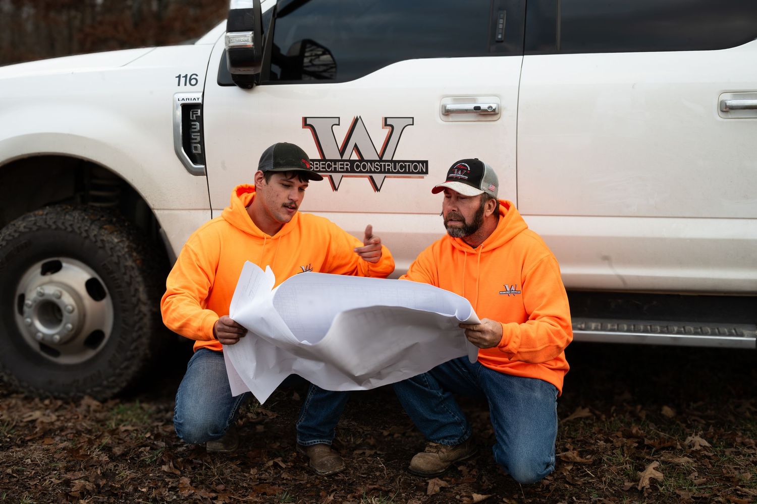 Workers in orange safety vests and jeans pose near white company trucks.