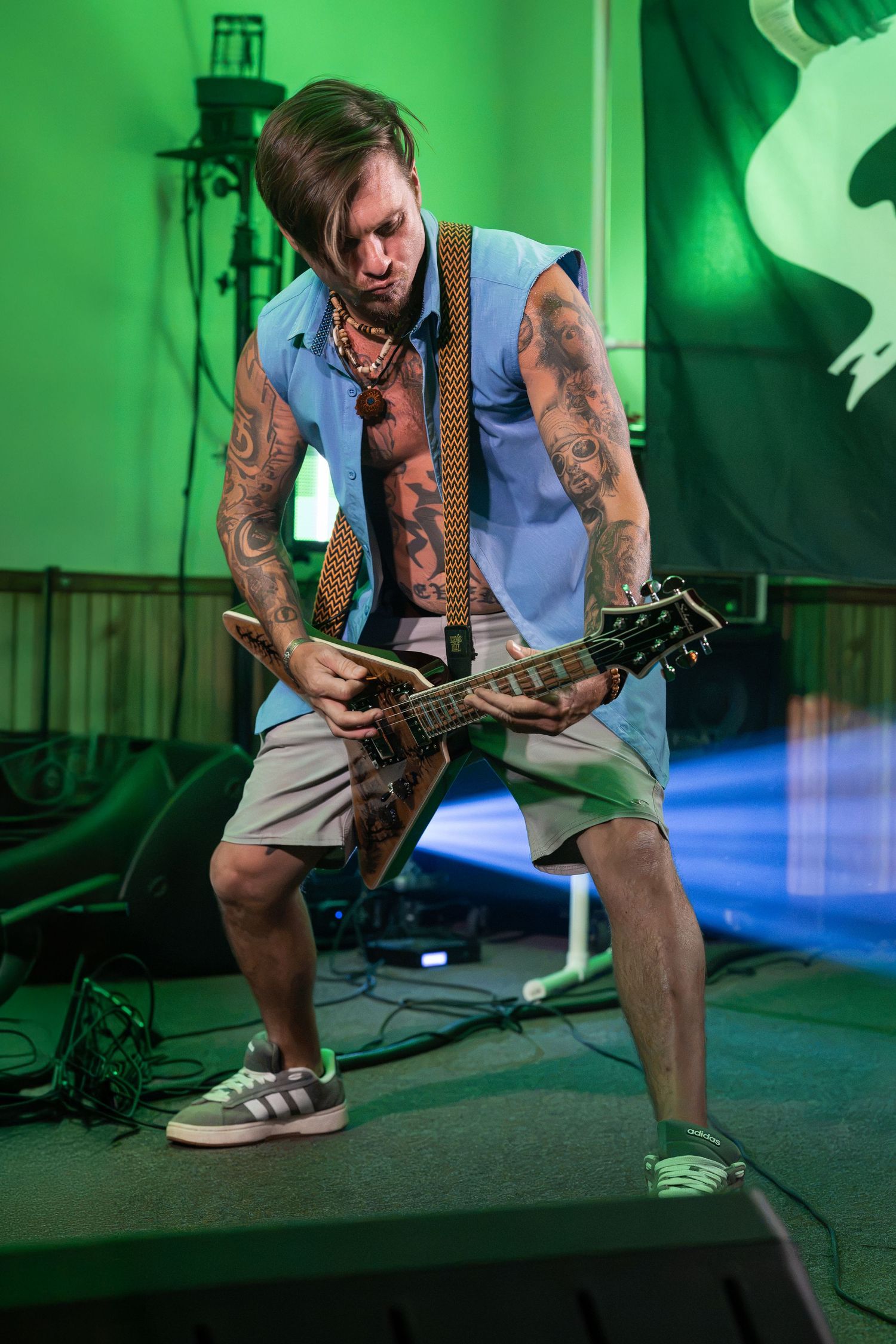 Tattooed male guitarist performs on stage with green lighting, playing electric guitar in sleeveless shirt and shorts.
