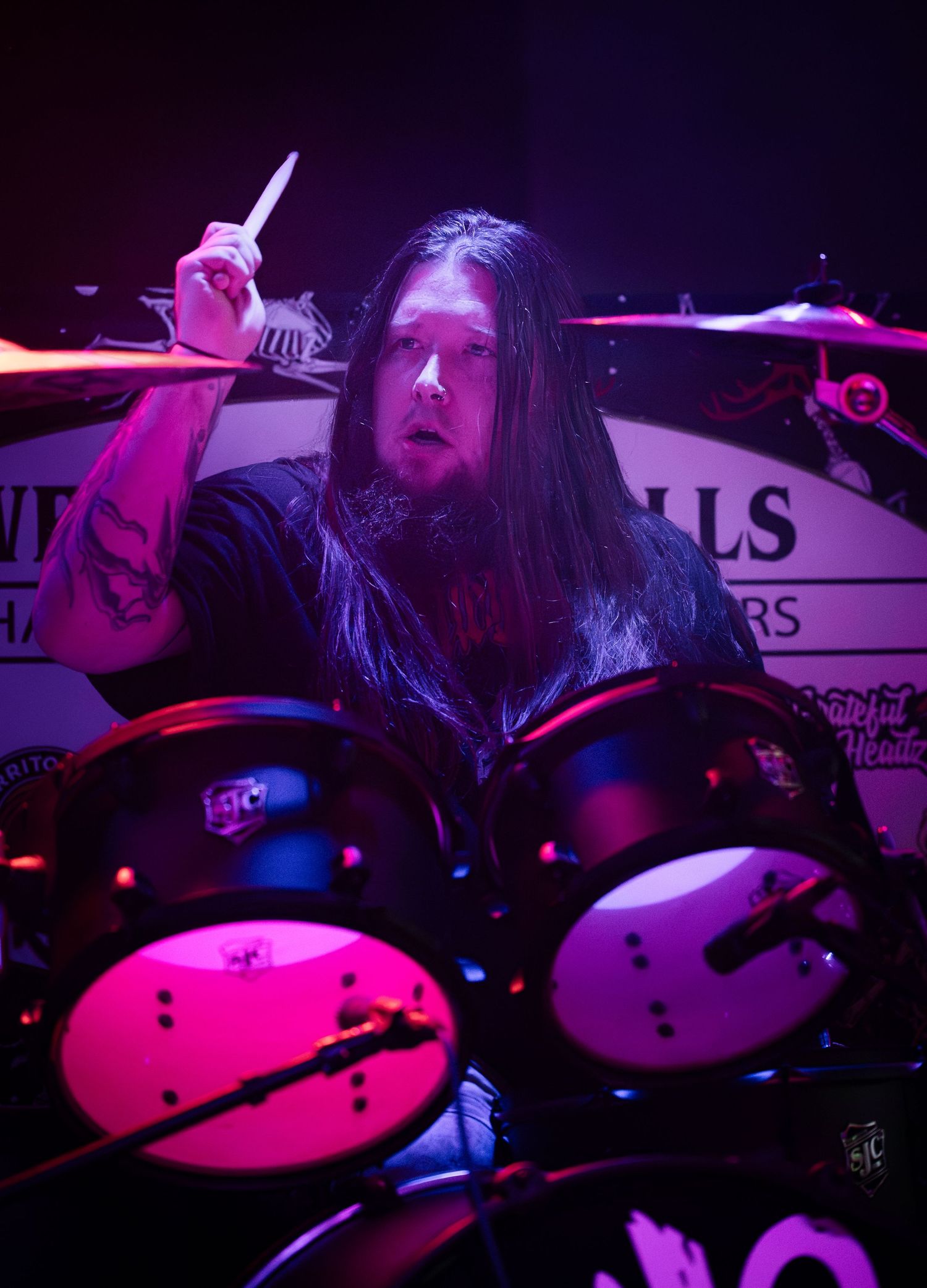 A drummer performs on stage illuminated by vibrant pink and purple lighting during a live music performance.