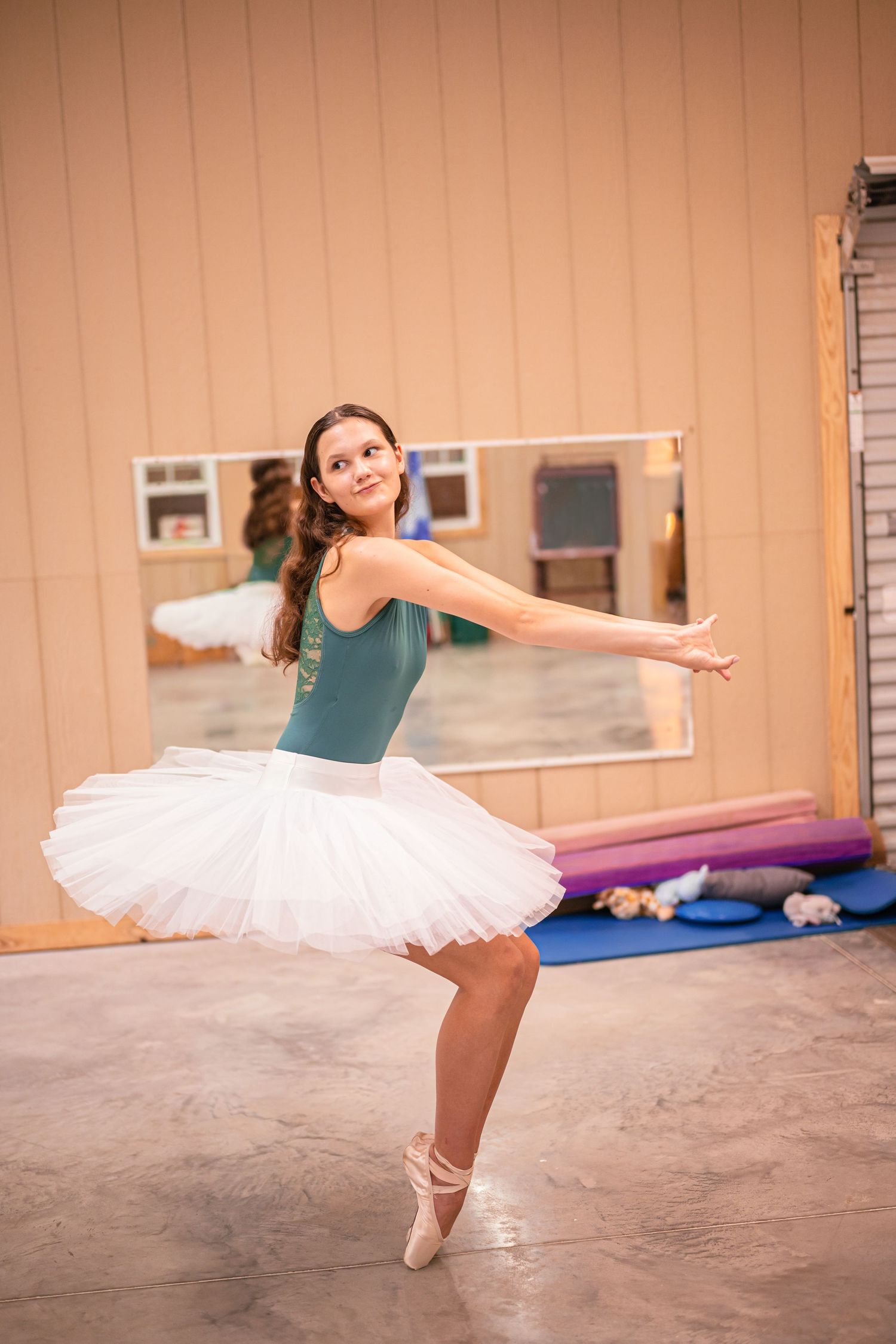Young ballet dancer in white tutu and teal leotard practices graceful poses in a dance studio.