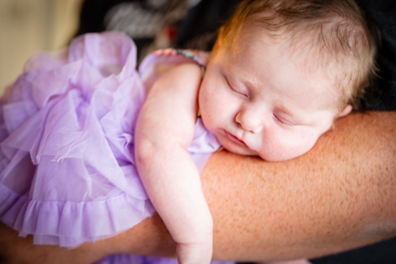 A sleeping baby wearing a purple tutu dress rests peacefully on an adult's arm.