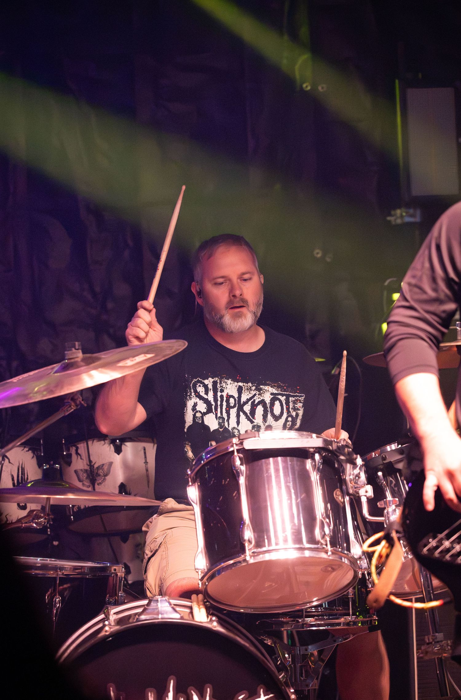 Drummer performs passionately behind a silver drum kit on stage with dramatic lighting effects.