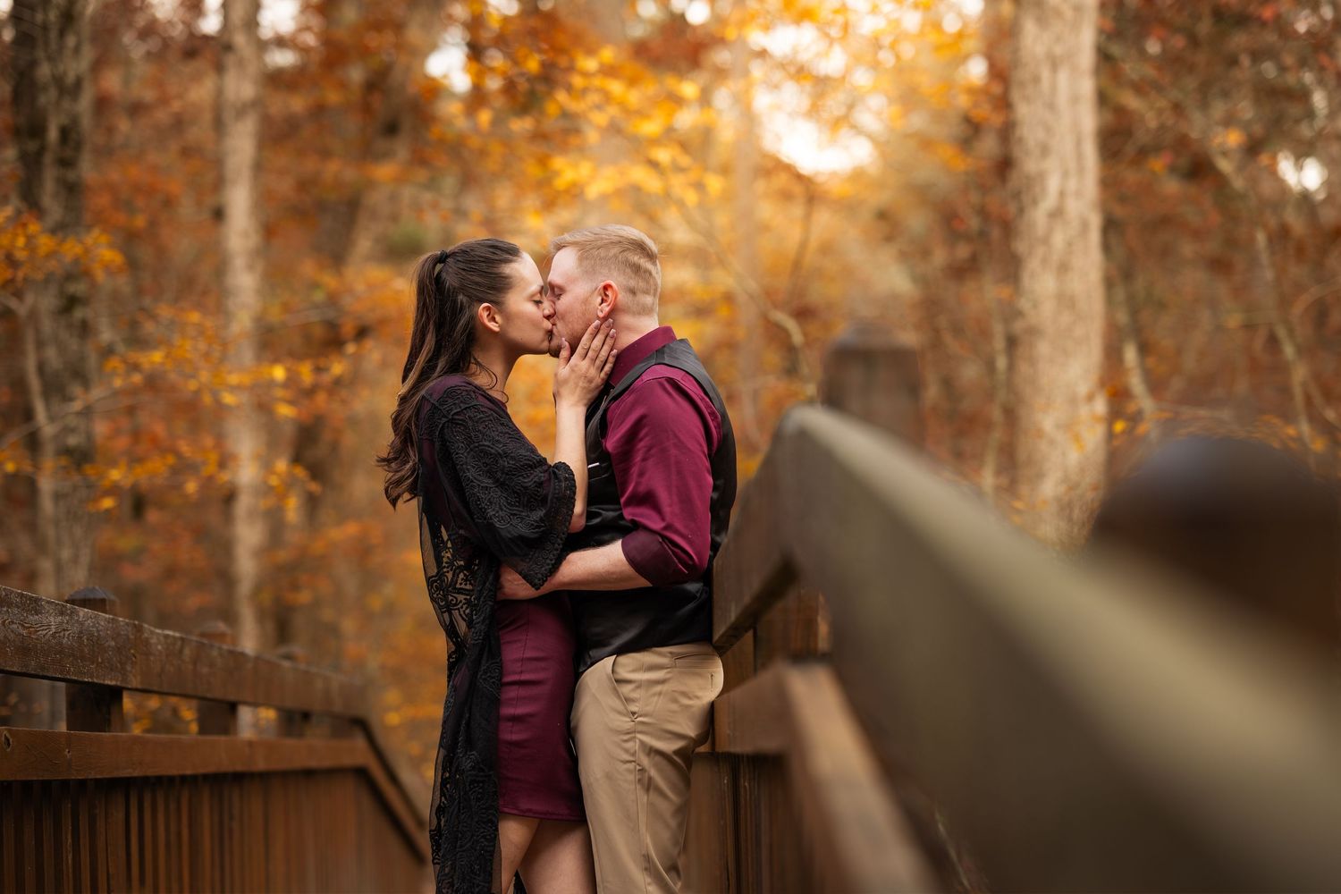 A romantic couple shares a tender moment on a wooden bridge surrounded by autumn foliage at sunset.