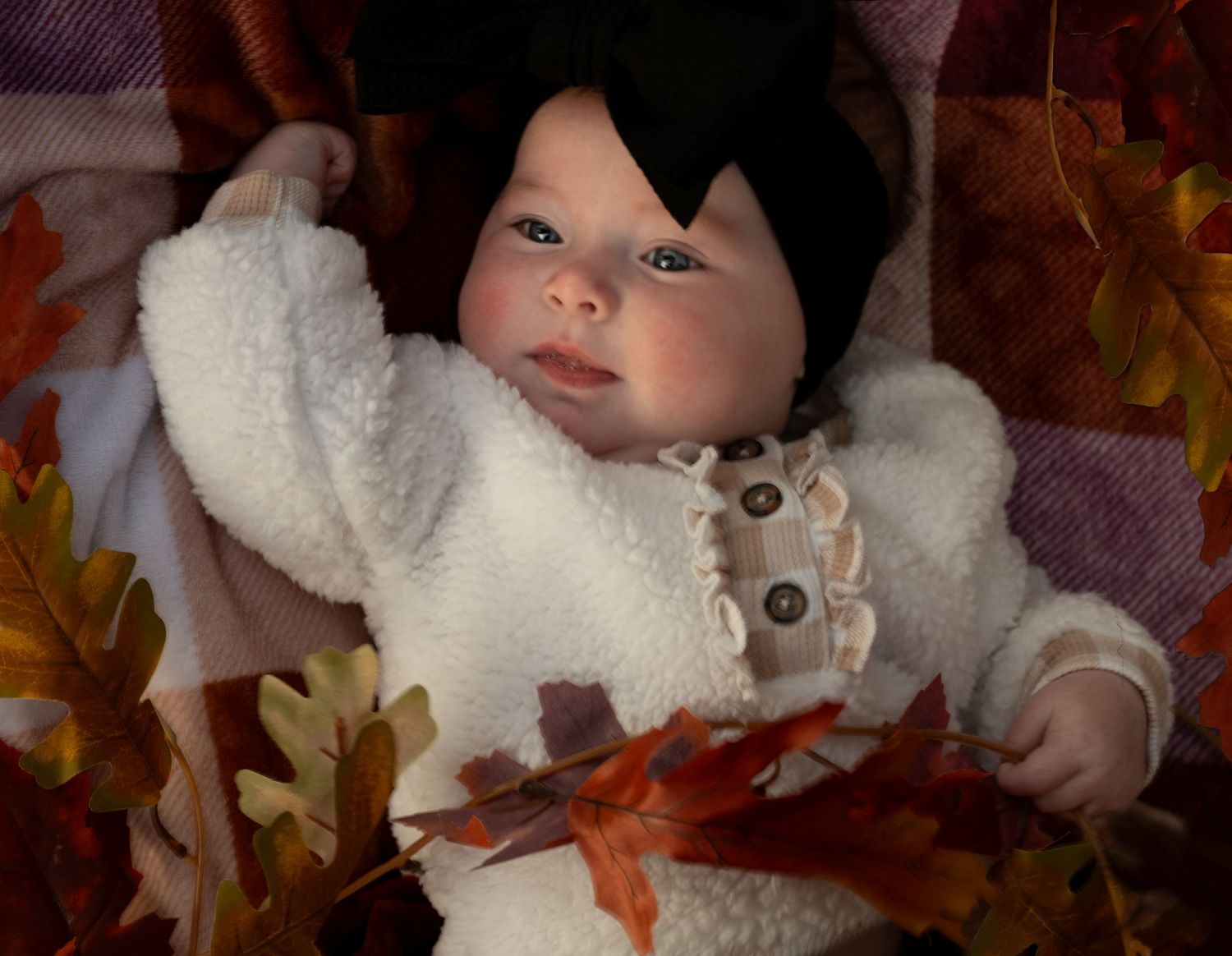 Baby wearing cozy white winter outfit lying among autumn leaves and fall decorations.