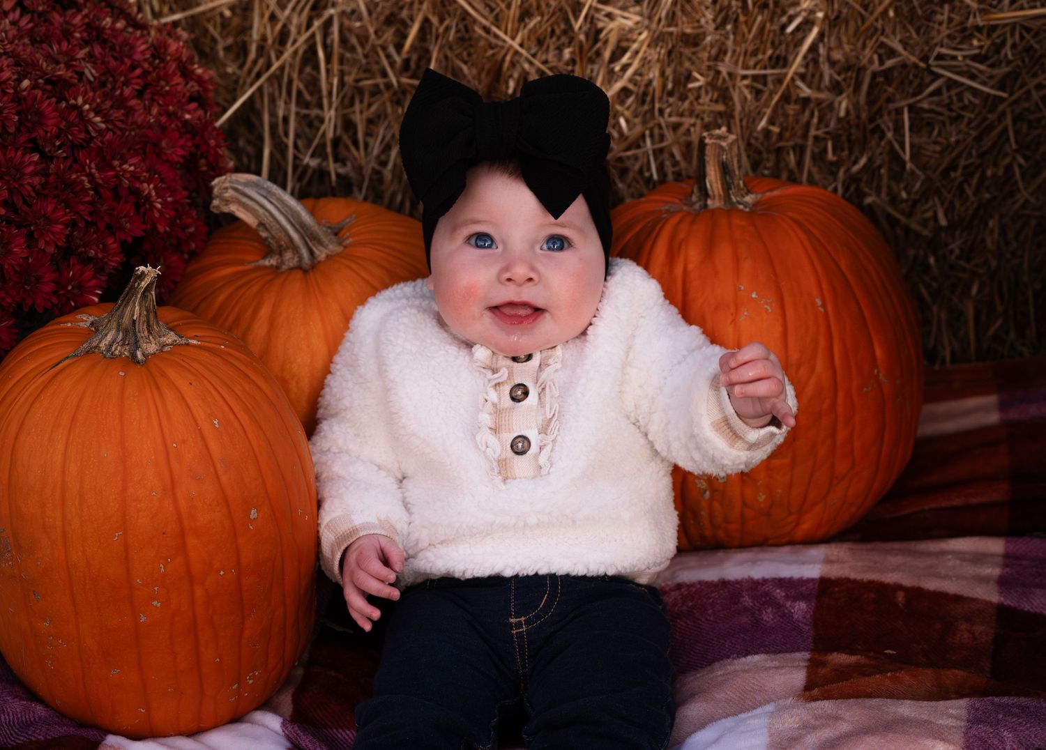A small figure in a white sweater and black bow sits between large orange pumpkins on hay bales for a fall photo.
