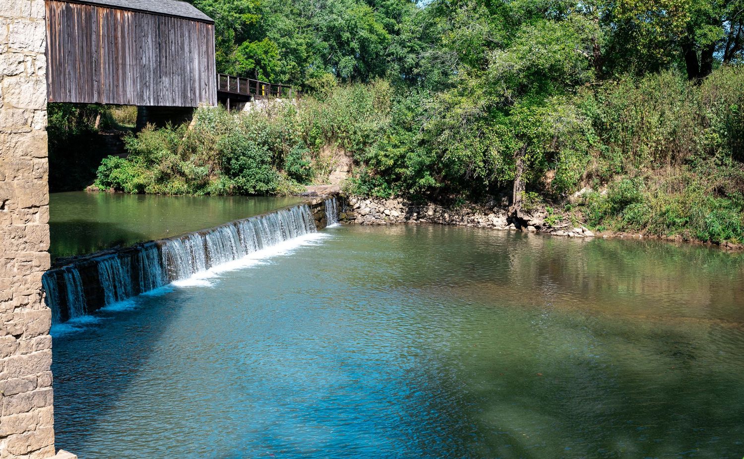 A serene waterfall cascades over a stone dam into a tranquil blue-green pool surrounded by lush vegetation.