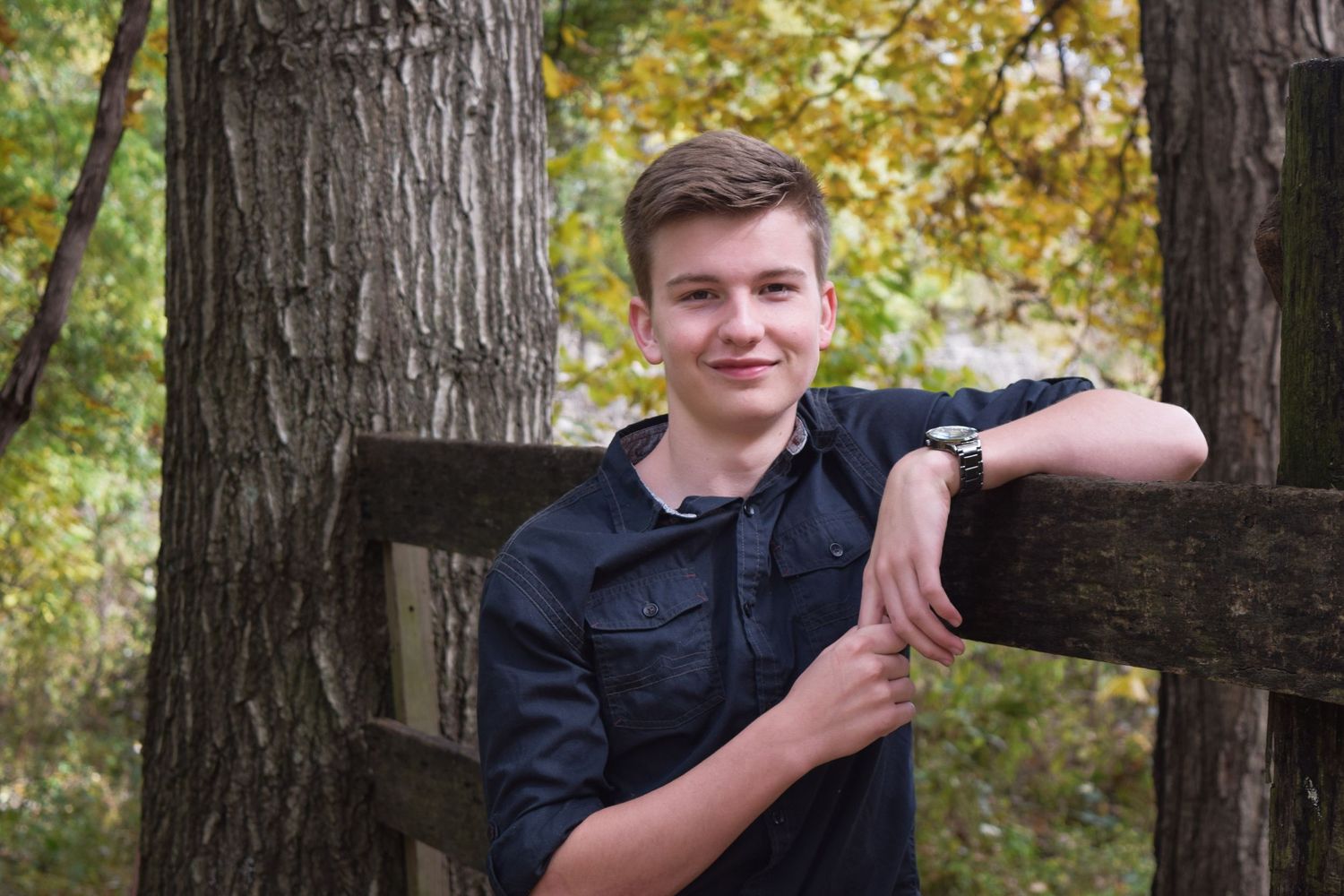 A person in a navy jacket poses against tree trunks in an autumn forest setting.